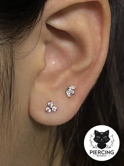 Close-up of a person's ear with two small, flower-shaped diamond stud earrings and a black logo featuring a cat's face and the text "Piercing Remedy" in the bottom right corner.