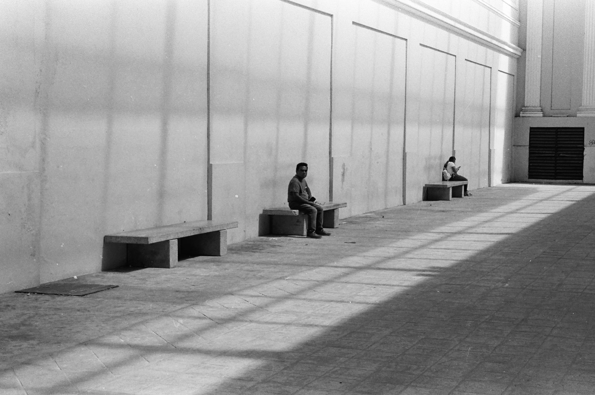 Black and white clinical composition of a man sitting alone on a concrete bench in a vast hallway. 35 mm black and white film.