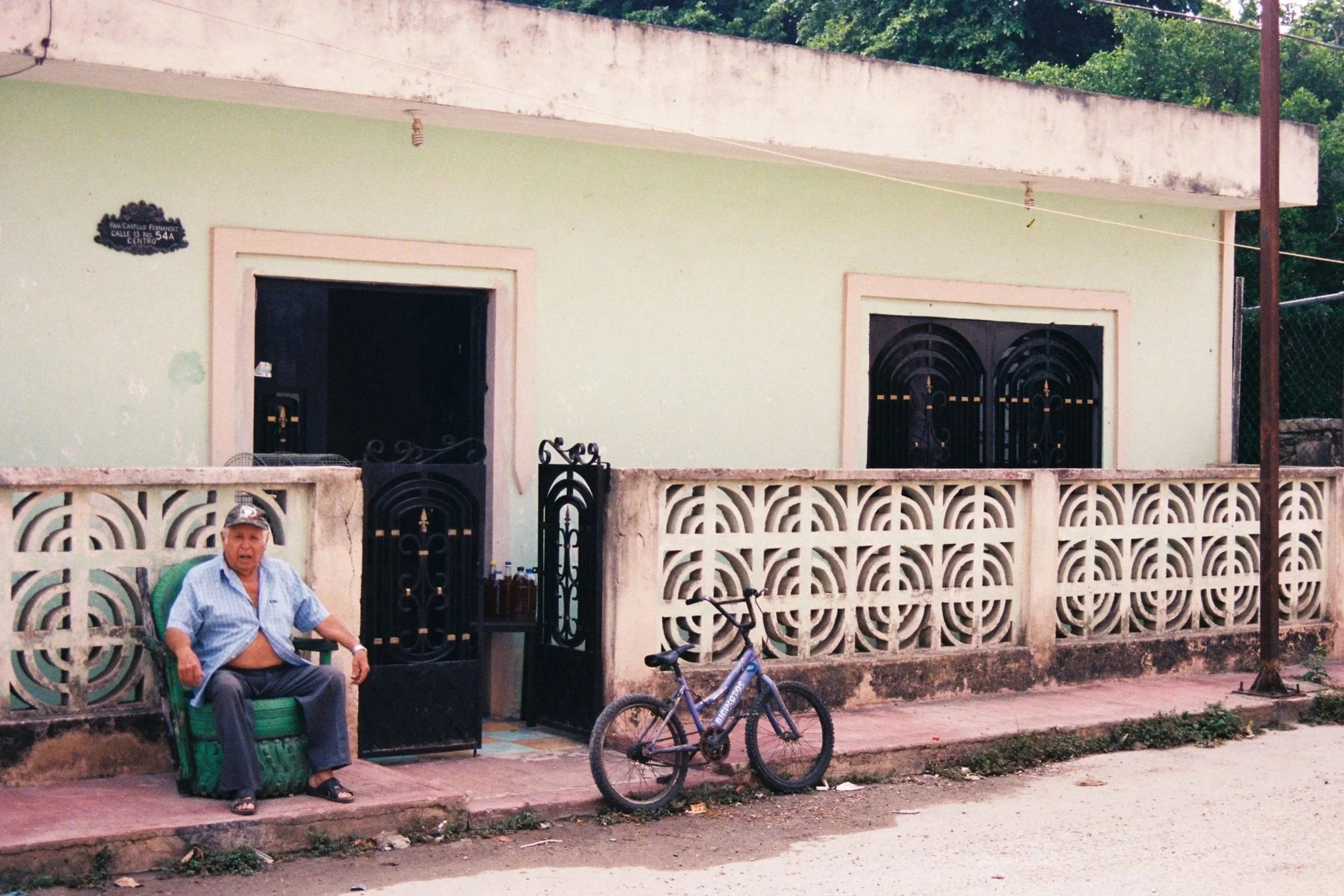 Elderly man sitting on a chair made of recycled tires in front of a lime green house facade, Mexico. 35 mm color film.