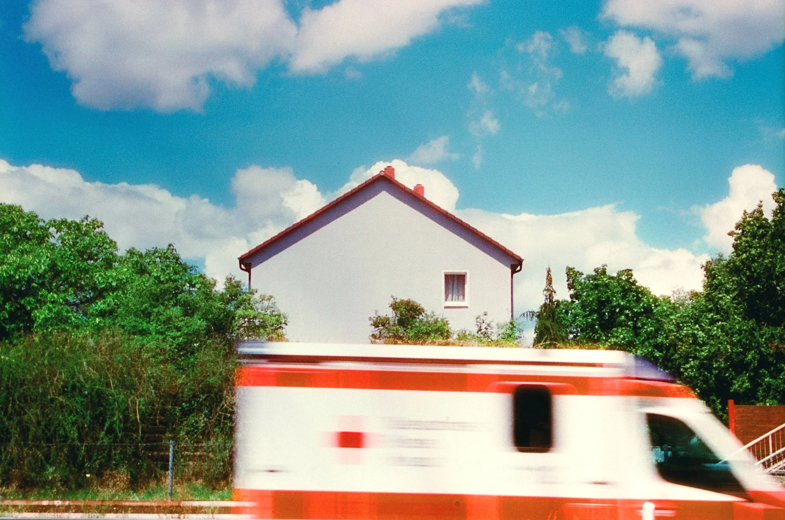A long-exposure shot of a suburban house with a red-roof gable; a blurred white ambulance with a red stripe and cross streaks across the foreground, creating a sharp contrast between domestic stillness and medical urgency. 35 mm color film.