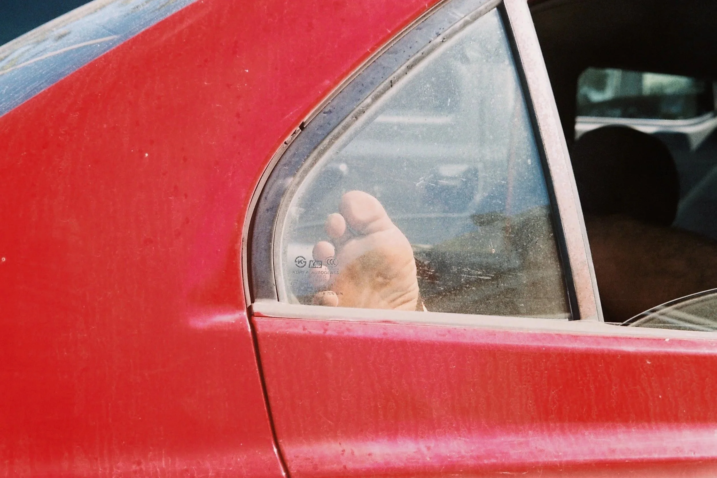 A close-up of a red car’s rear window; behind the dusty, sunlit glass, a bare human foot is visible, resting against the pane, isolating the limb from the rest of the body. 35 mm color film.