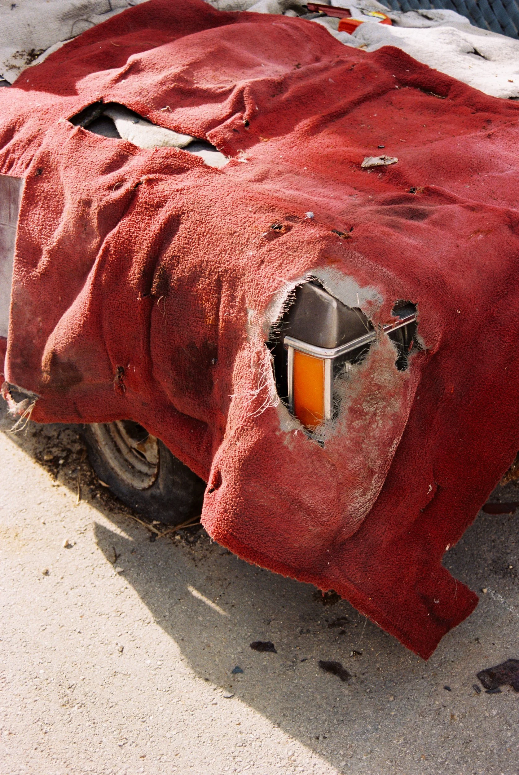 A close-up of a vehicle covered by a heavy, tattered red carpet; the carpet is torn, exposing a single orange turn signal light and rusted metal beneath the fabric. 35 mm color film.