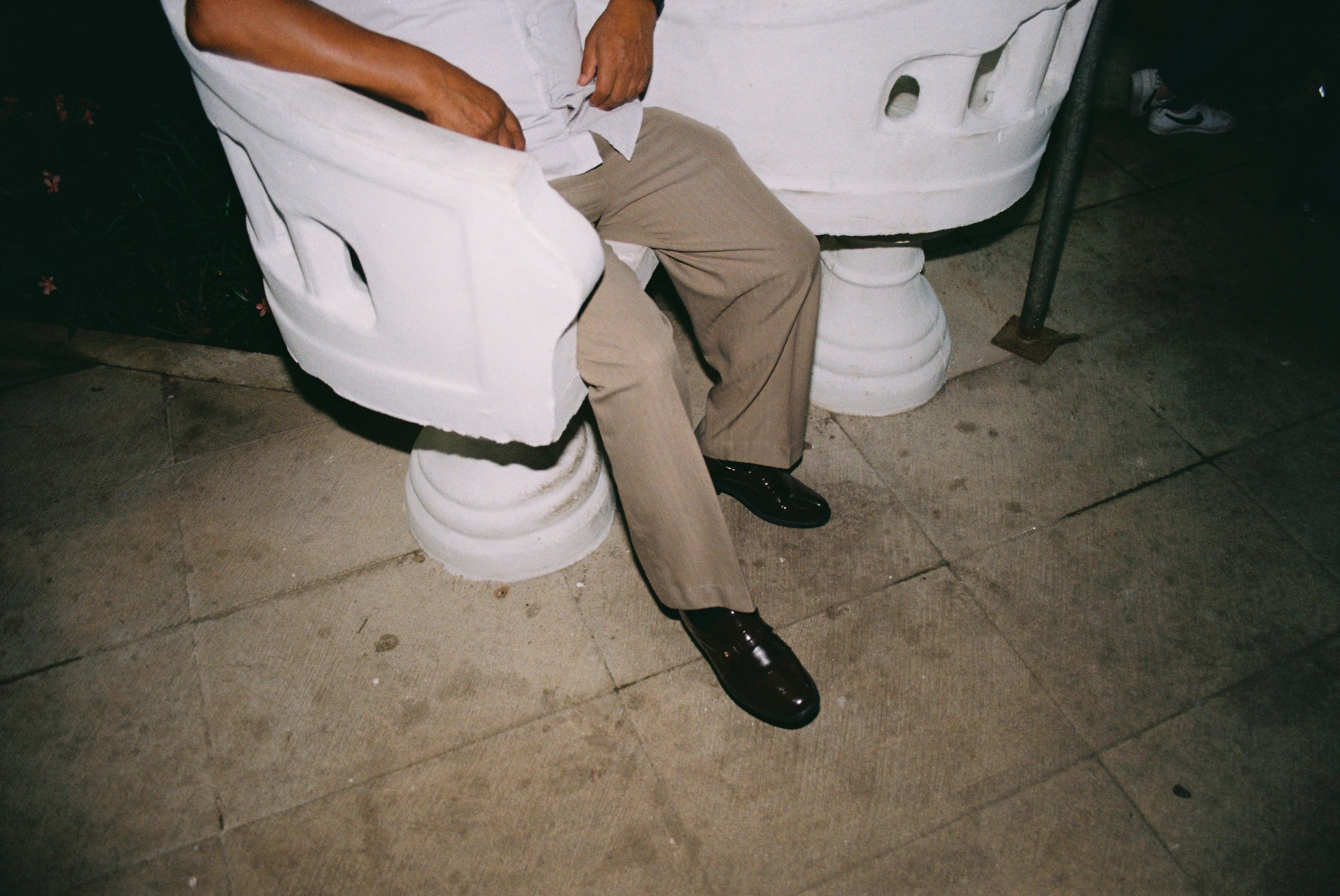 Minimalist detail of a man's hands and legs seated in a white concrete public chair. 35 mm color film.
