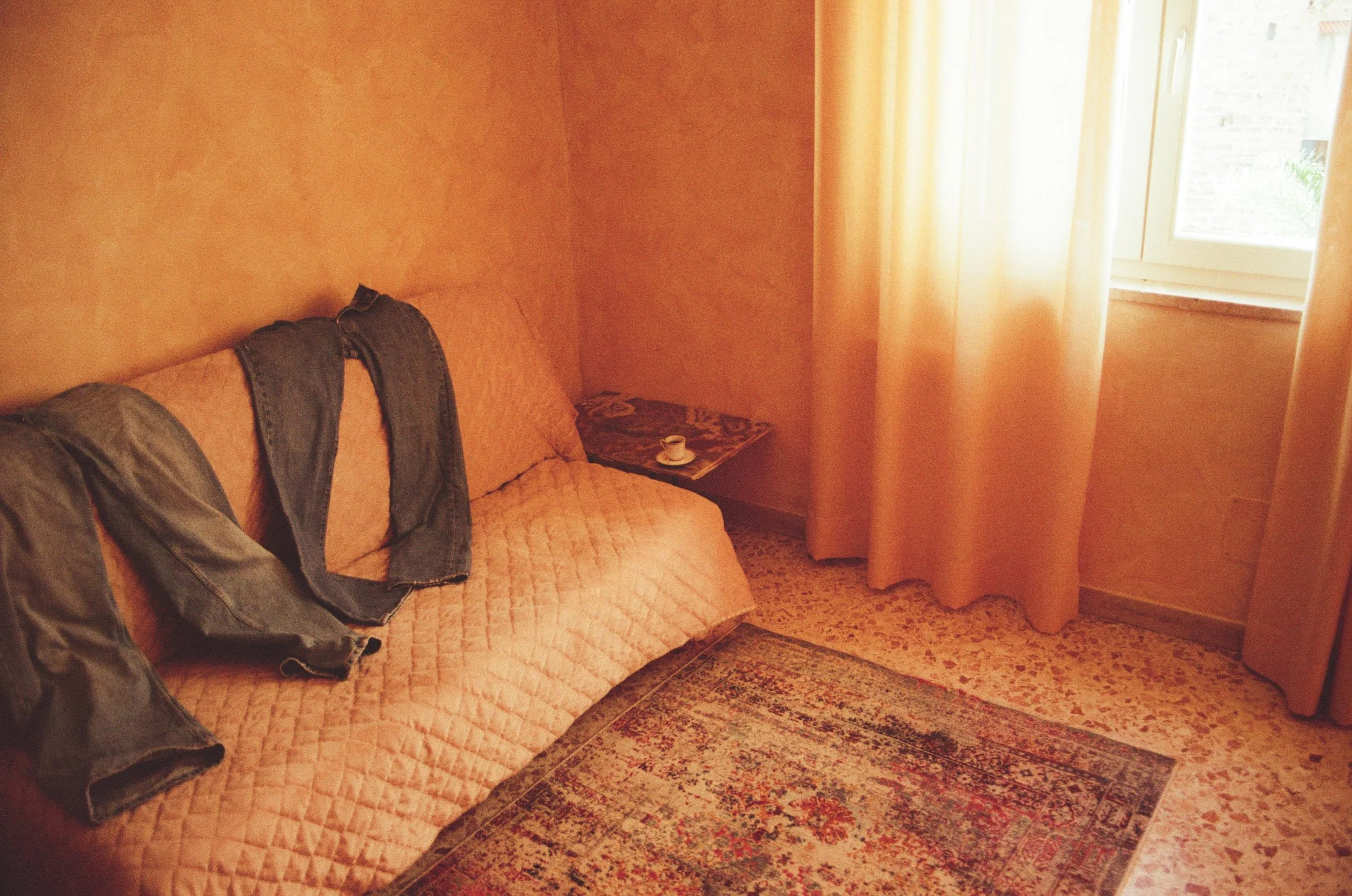 Blue jeans draped over a peach sofa in a sun-filled room, a warm but bitter image of a hollowed-out domestic space. 35 mm color film.