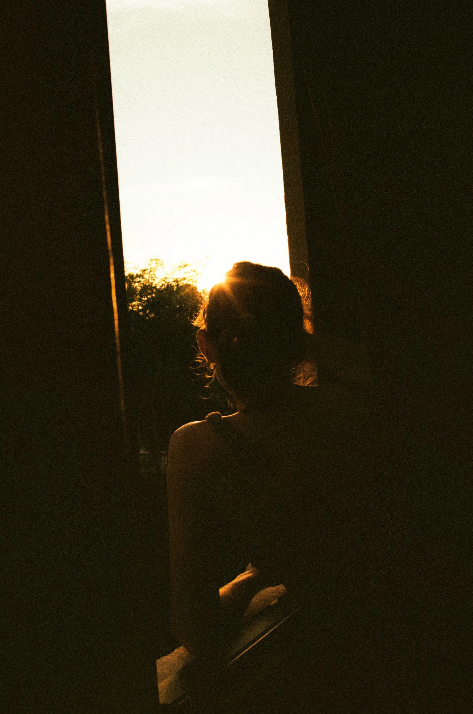 Silhouette of a person framed by a glowing window at golden hour, a melancholic observation of light and memory. 35 mm color film.