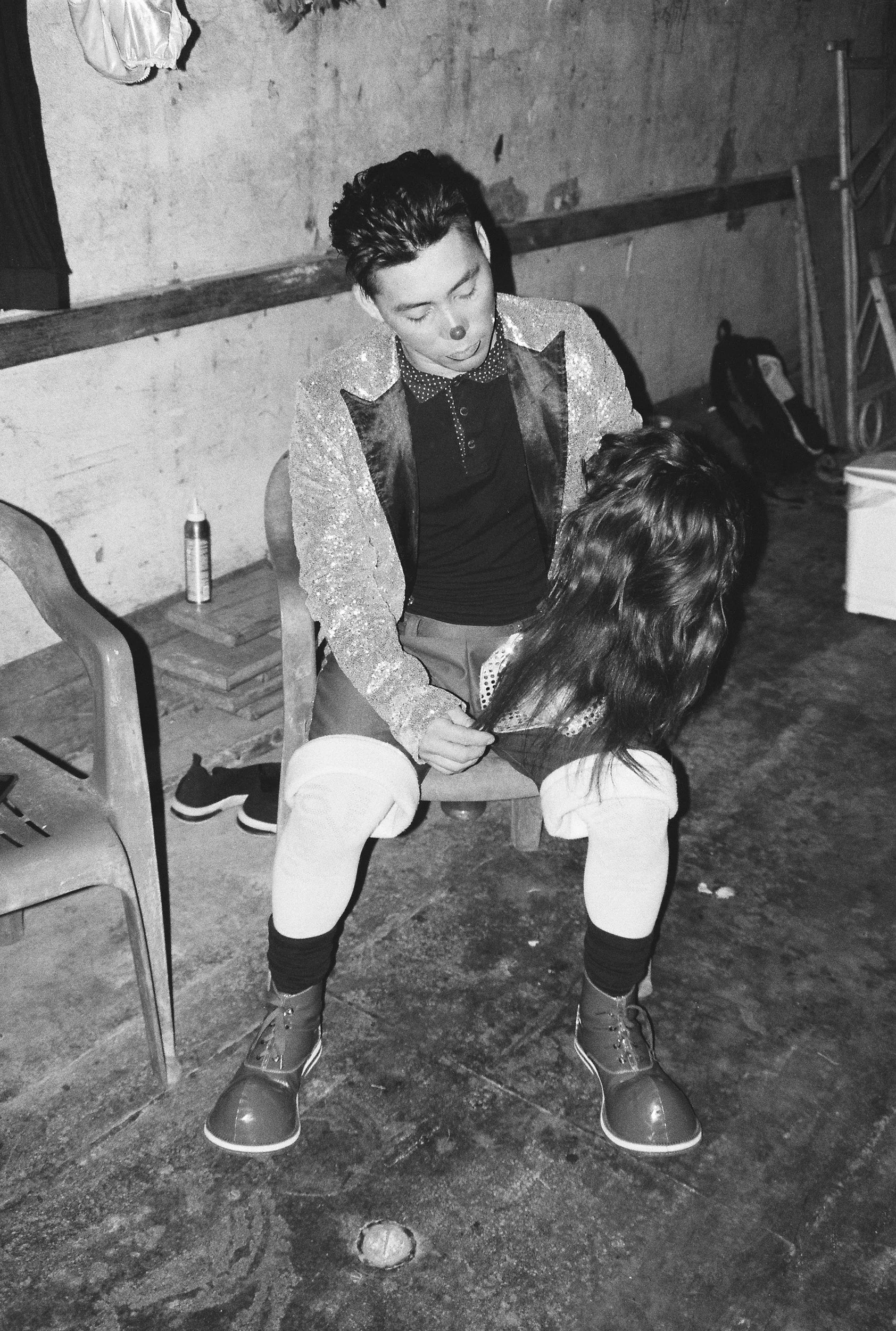 Analog portrait of an exhausted clown sitting in a dark room, capturing the tragedy of Mexican street performance. 35 mm black and white film.