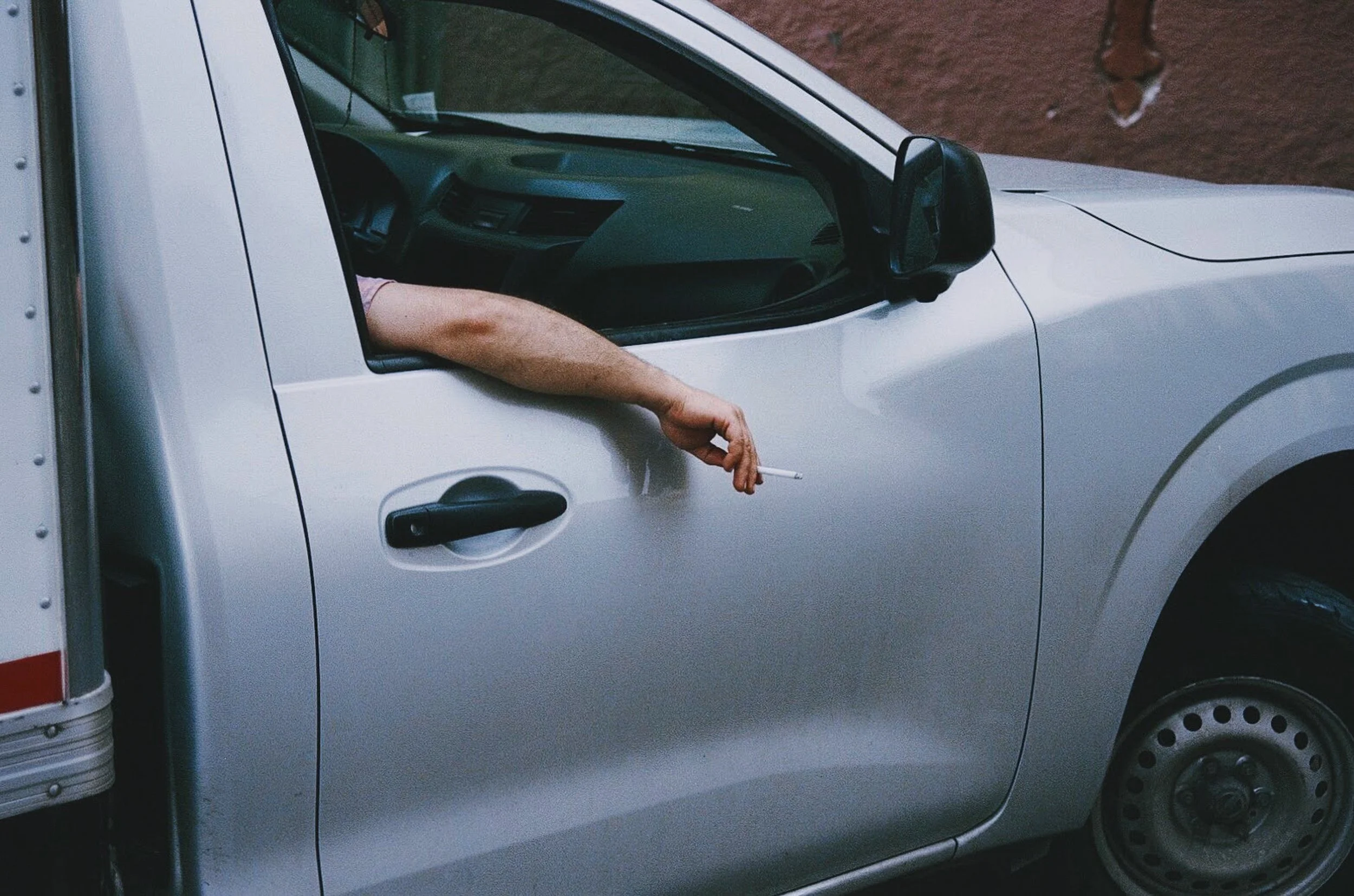 Man's arm resting on a white truck window holding a lit cigarette, representing waiting and boredom. 35 mm color film.