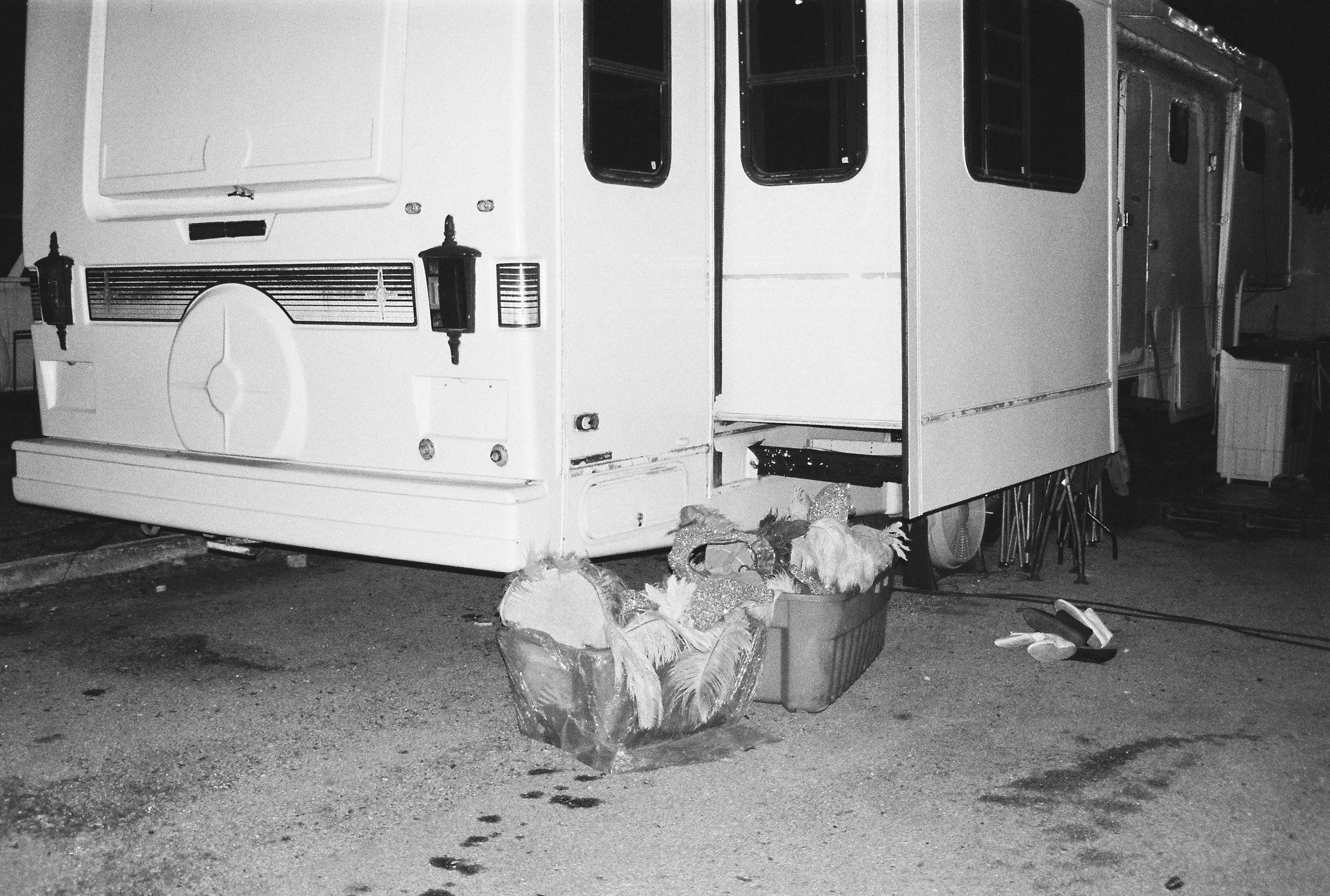 Analog photograph of  large camper at night, with a plastic bin full of show feathers and sequins on the ground, representing the raw backstage of Mexican entertainment. 35 mm black and white film. 
