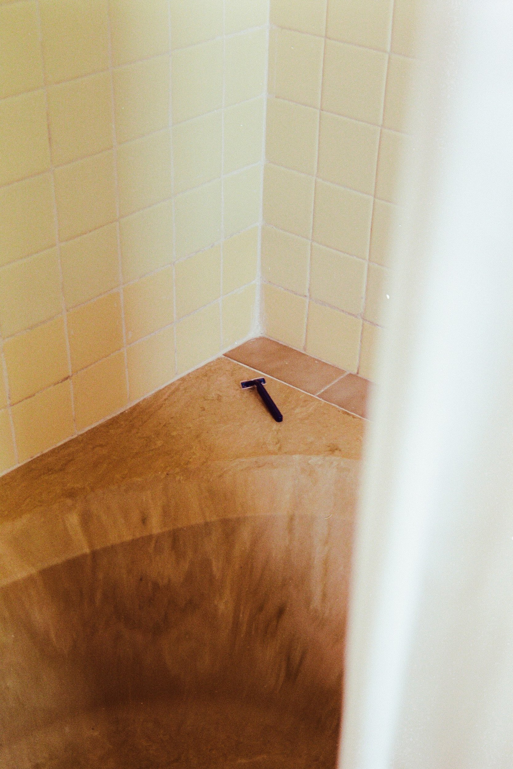 A solitary blue razor left on the edge of a tan-tiled bathtub, a clinical trace of a departed presence. 35 mm color film.