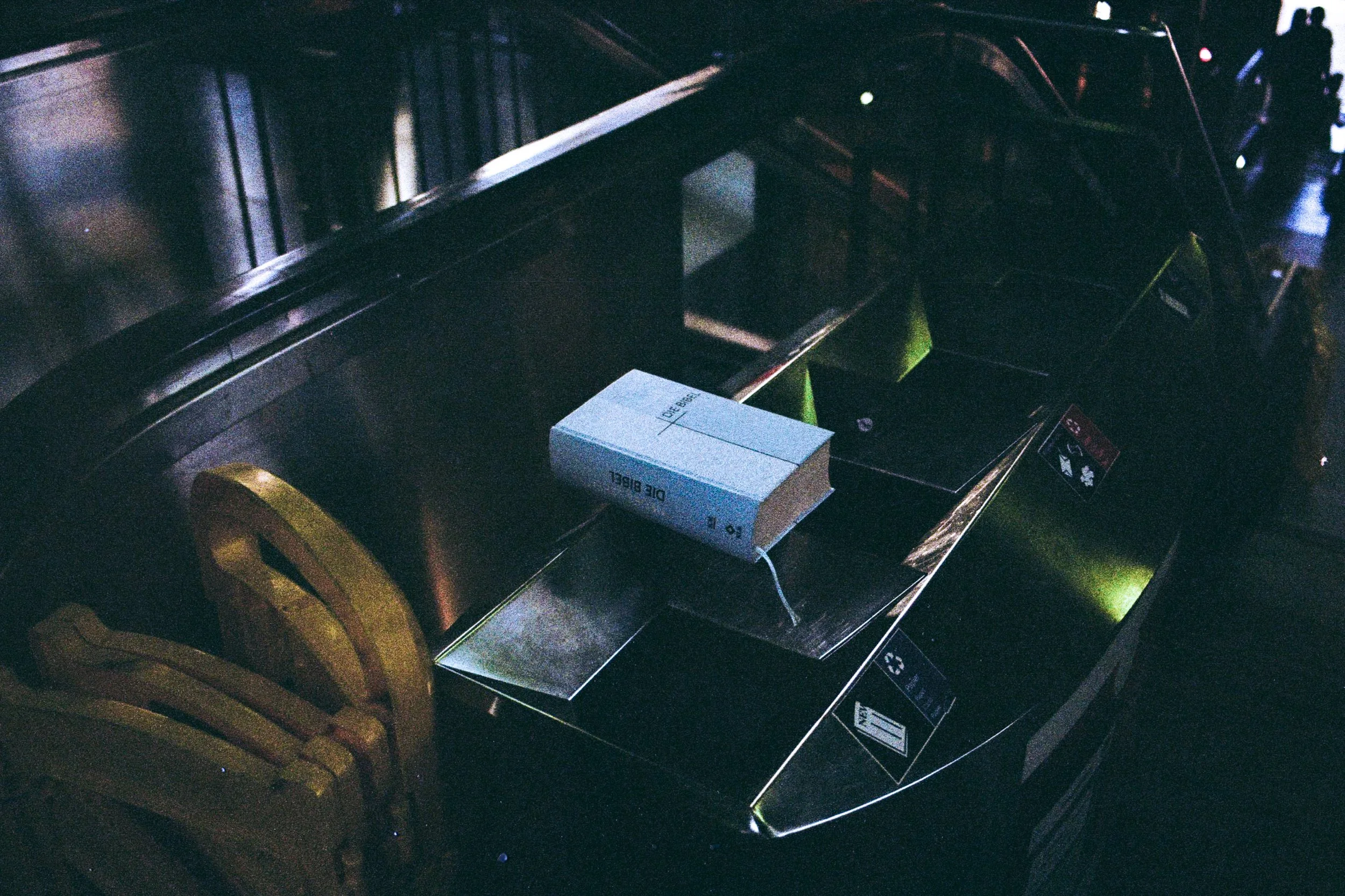 A blue hardcover Bible titled "Die Bibel" sits abandoned on the flat, brushed metal top of a public trash can. The book is positioned in a dark, industrial environment near the metallic structures of an escalator, marking a stark contrast between rel