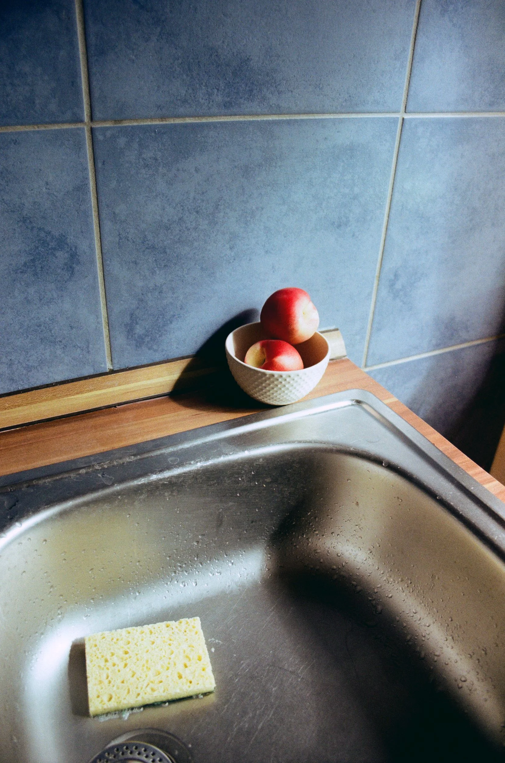 Still life of red peaches in a white bowl by a kitchen sink, where domestic warmth meets a quiet, bittersweet solitude. 35 mm color film.
