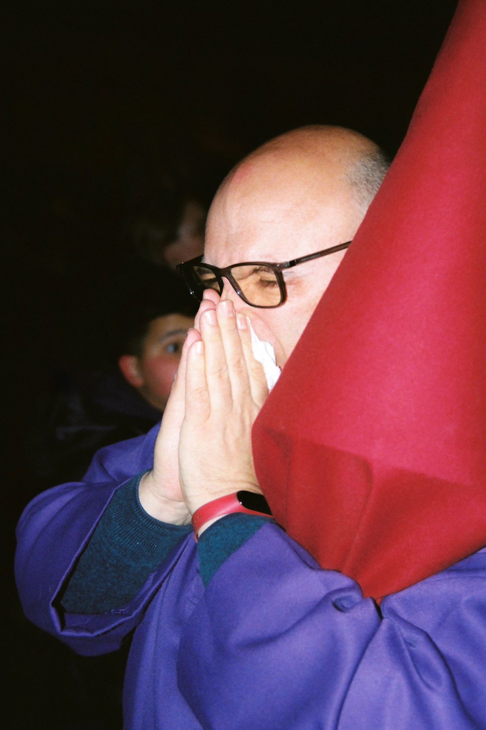 A person in purple ceremonial robes with their hands clasped over their face in an ambiguous gesture; a large, pointed red fabric cone (capirote) is positioned in the foreground, obscuring the side of their head. 35 mm color film.