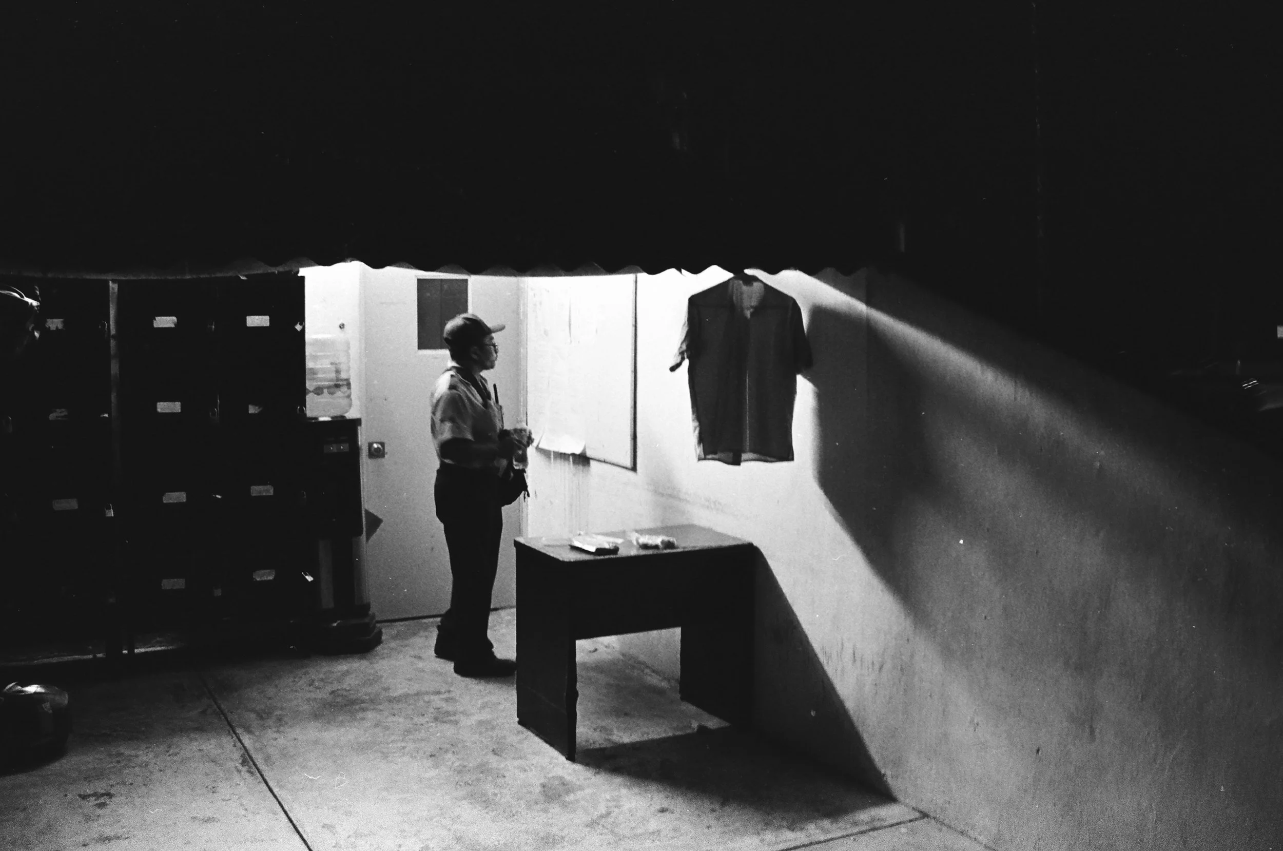 Night scene in black and white showing a security guard next to a shirt hanging under a harsh spotlight. 35 mm black and white film.