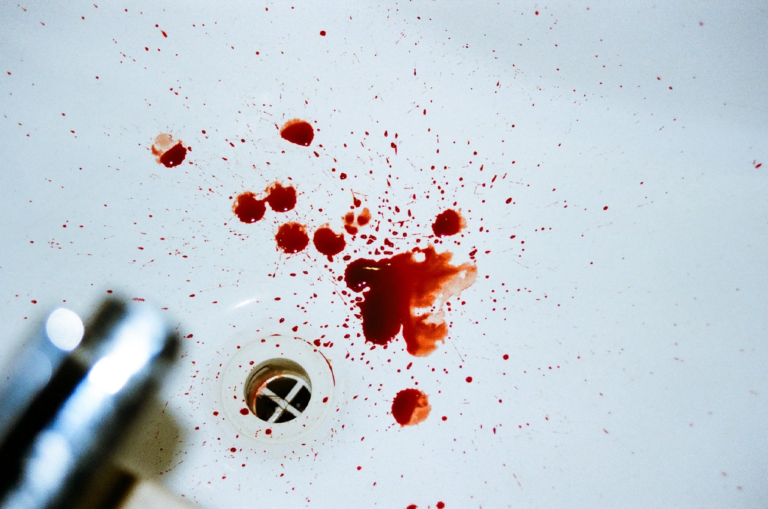A top-down view of a white ceramic sink basin with red blood splatters radiating from the center toward the drain; the chrome faucet is visible in the blurred foreground. 35 mm color film.