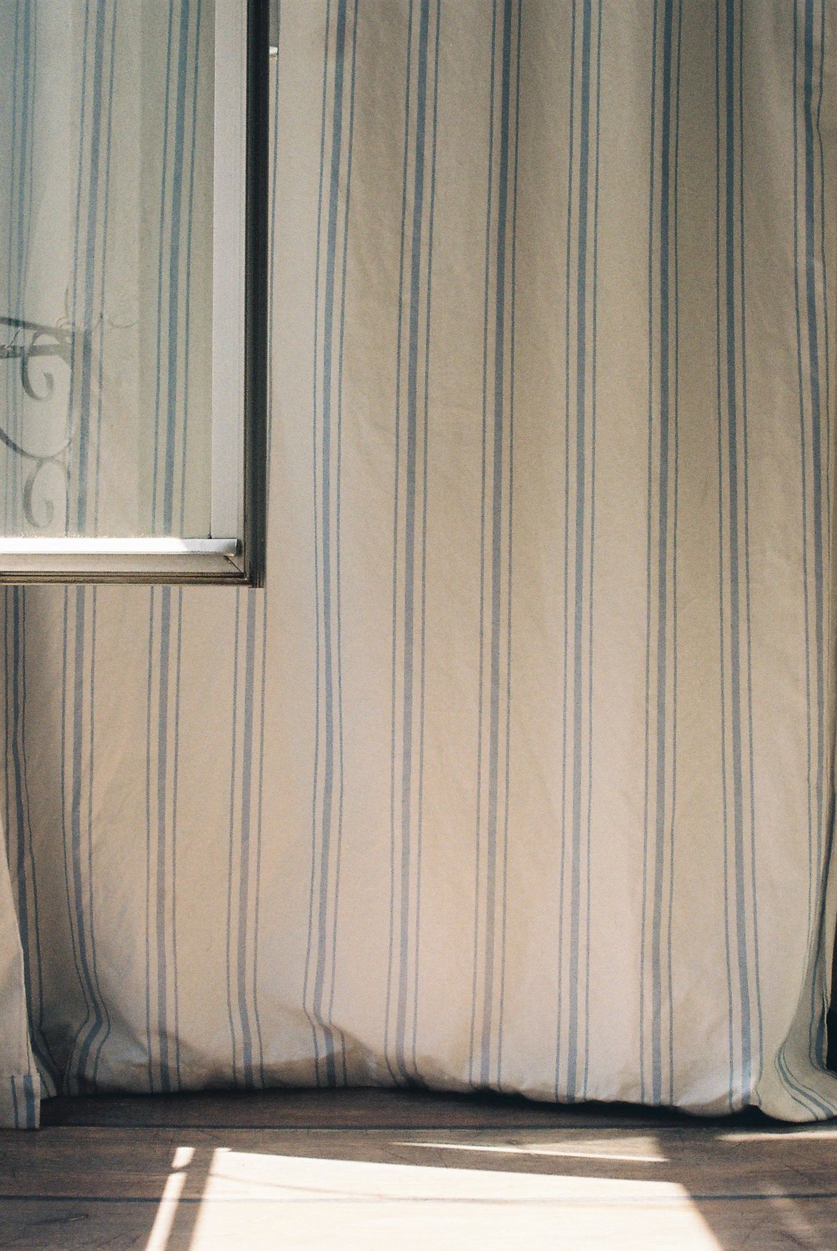 Soft natural light hitting a blue-striped curtain by an open window, symbolizing the threshold between shared and solitary space. 35 mm color film.