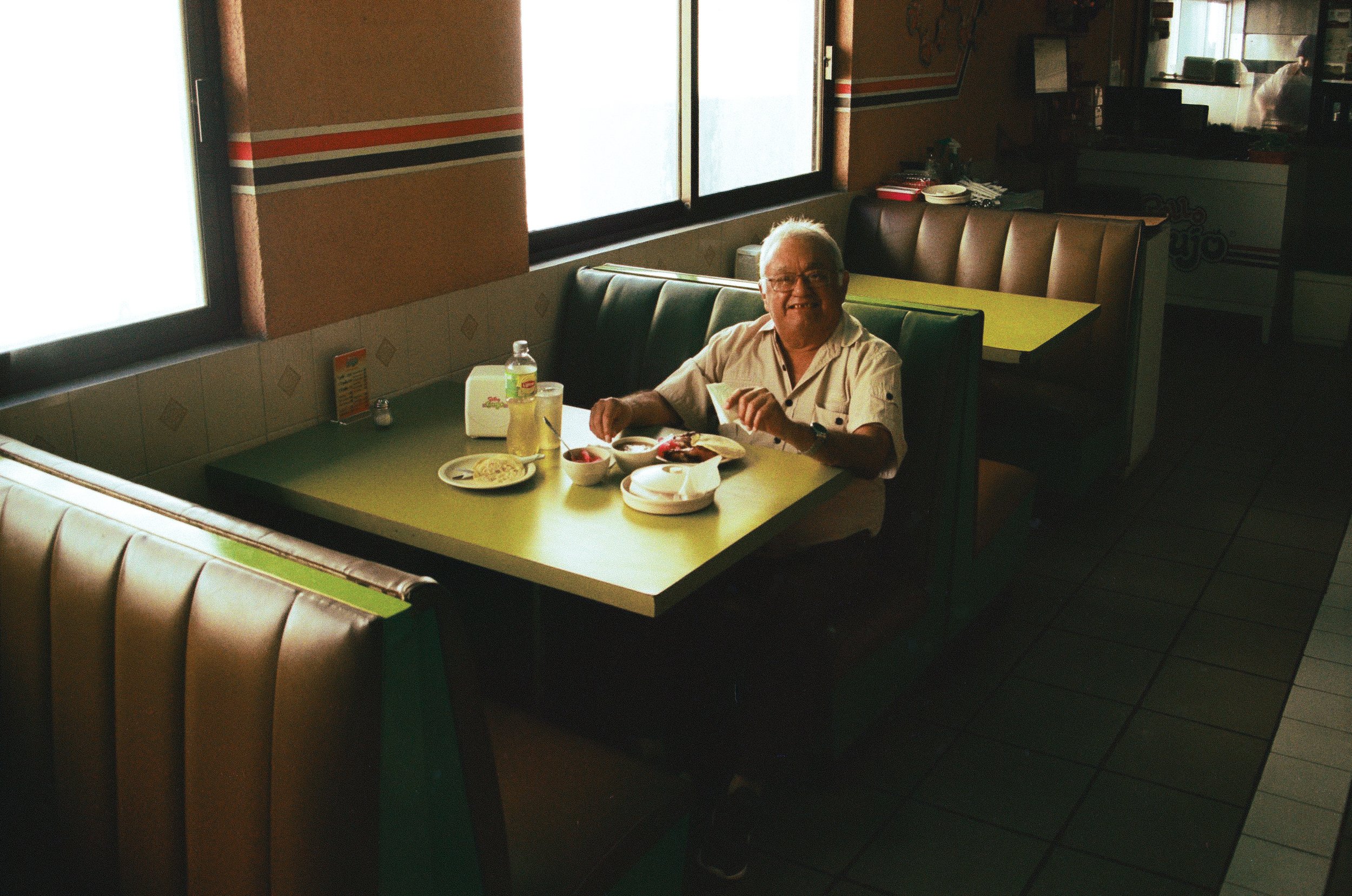 Smiling elderly man eating alone in a vintage diner booth, capturing the 'facade' of daily life. 35 mm color film.