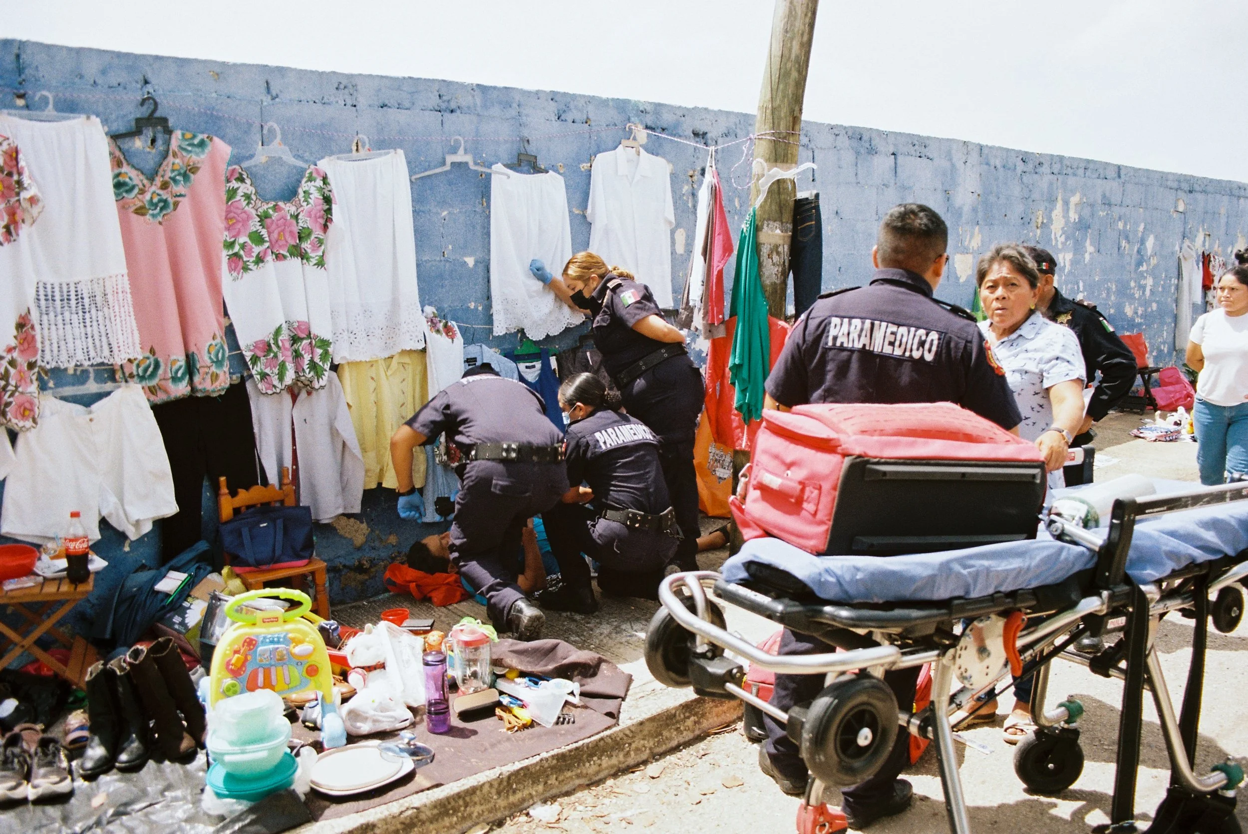 Paramedics attending to an individual at an outdoor market under hanging clothes, clinical realism. 35 mm color film.