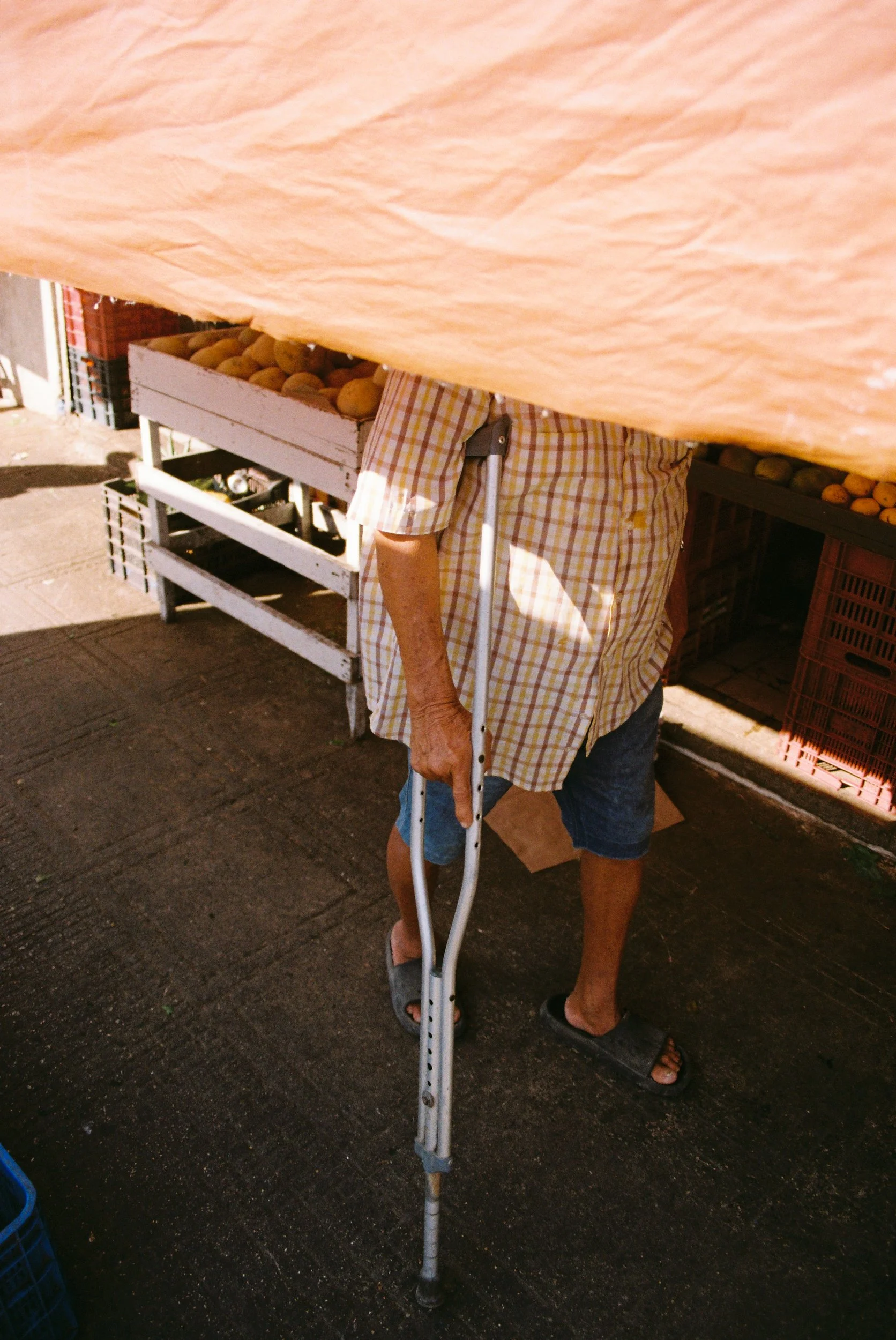 Back view of a man leaning on a metal crutch under a bright orange tarp, exploring physical vulnerability. 35 mm color film.