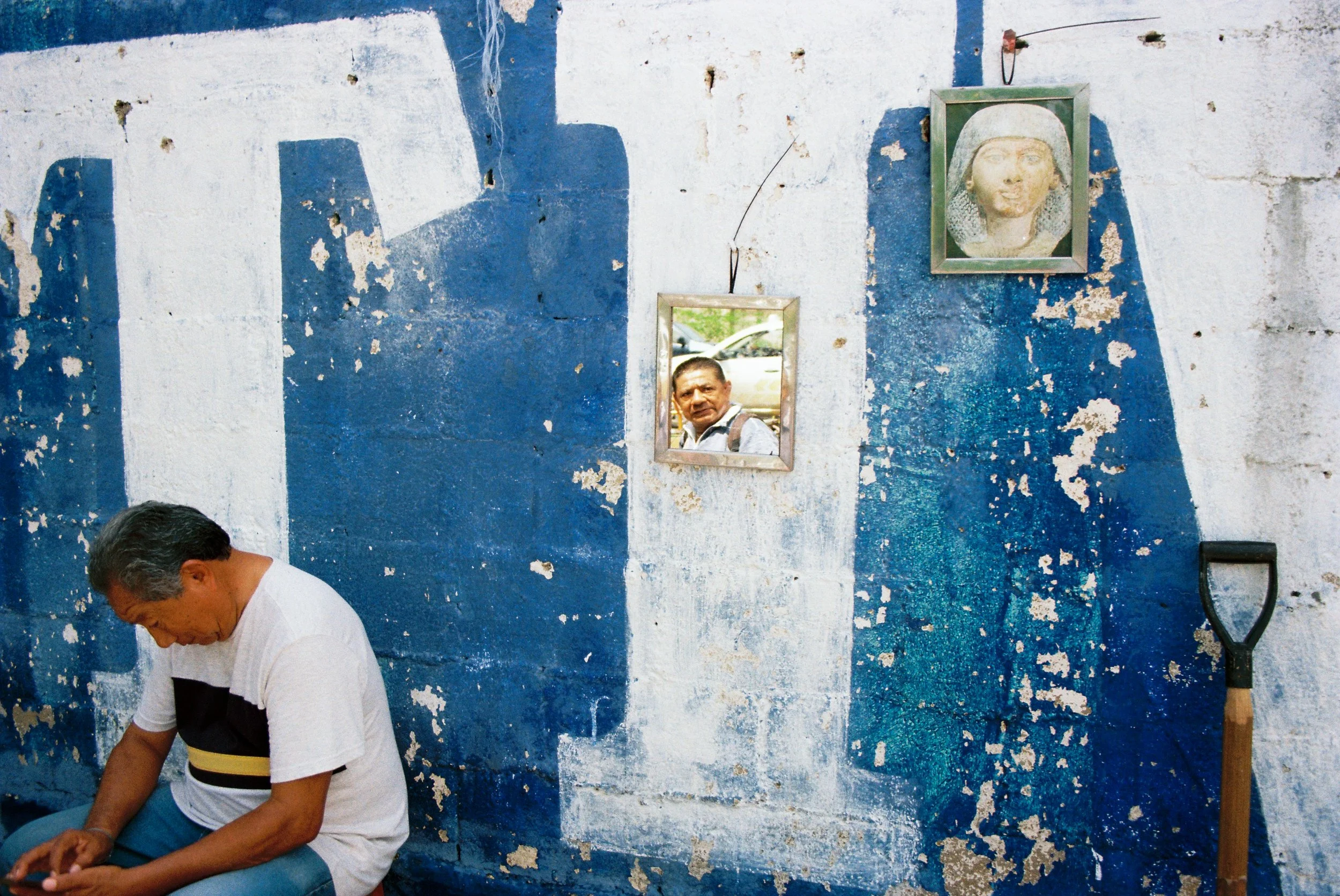 Small mirror hanging on a peeling white and blue wall reflecting a man's face, fragmented identity. 35 mm color film.