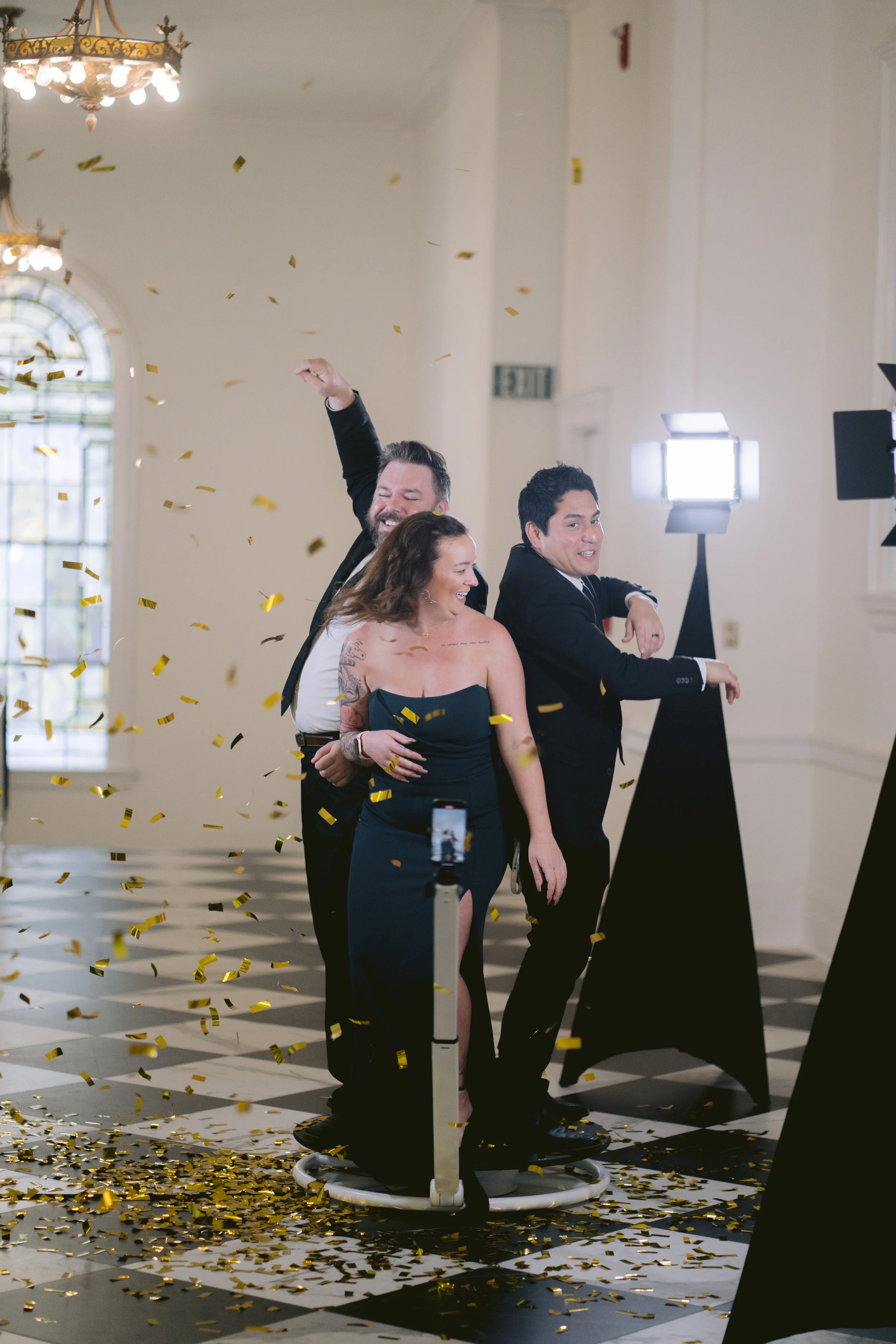 1 lady and 2 men dancing on a 360 photo booth in a hall with studio lights in the background and confetti on the floor 