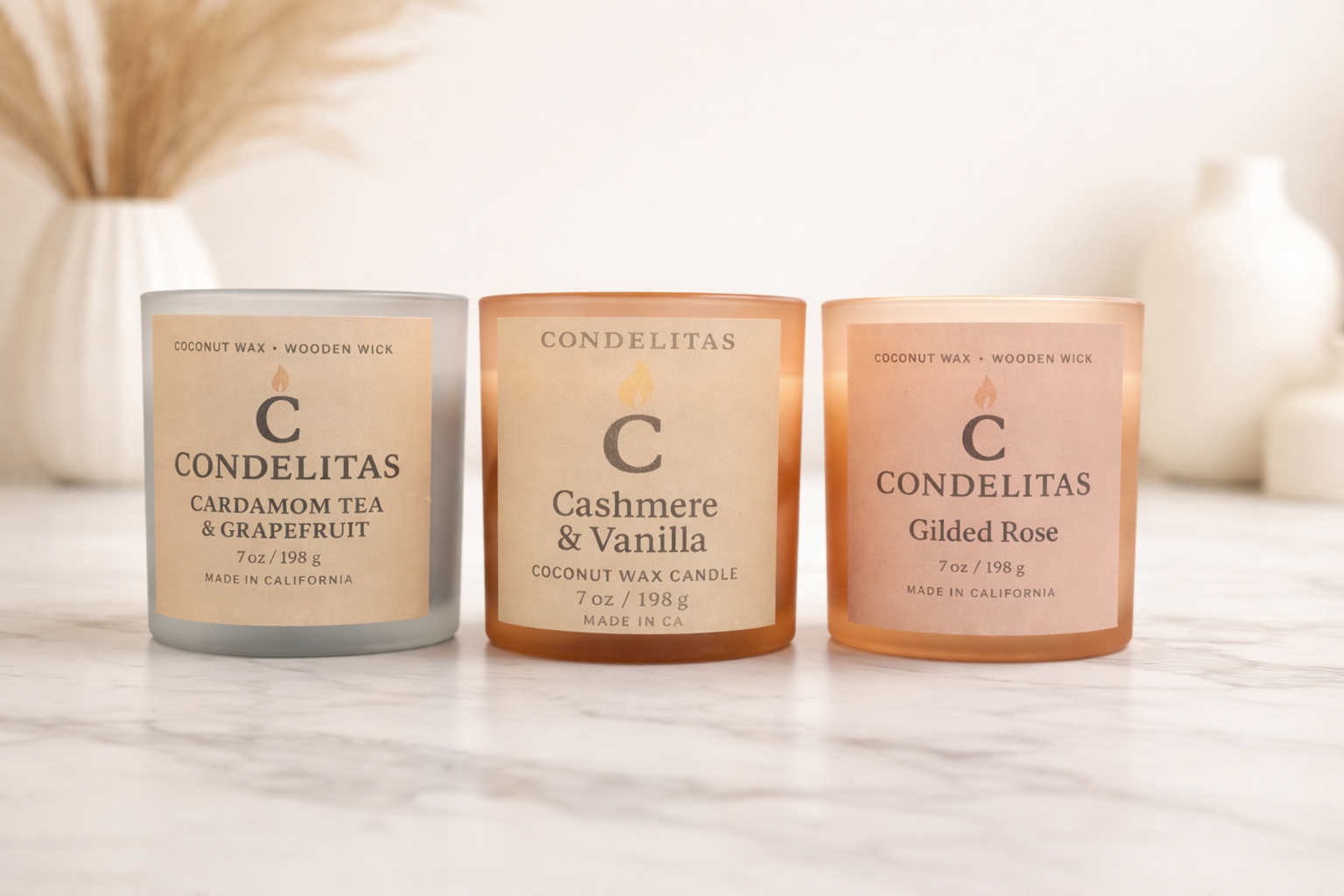 Three candles in frosted glass containers with minimalist labels on a marble surface, with vases in the background. The left candle is labeled 'Cardamom Tea & Grapefruit,' the middle 'Cashmere & Vanilla,' and the right 'Gilded Rose' by Condelitas.