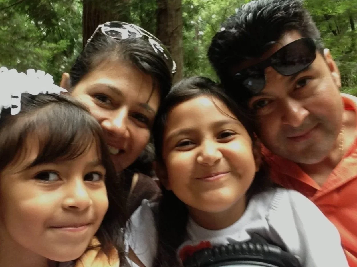 A group of five people, including children and adults smiling and taking a selfie outdoors with trees in the background.