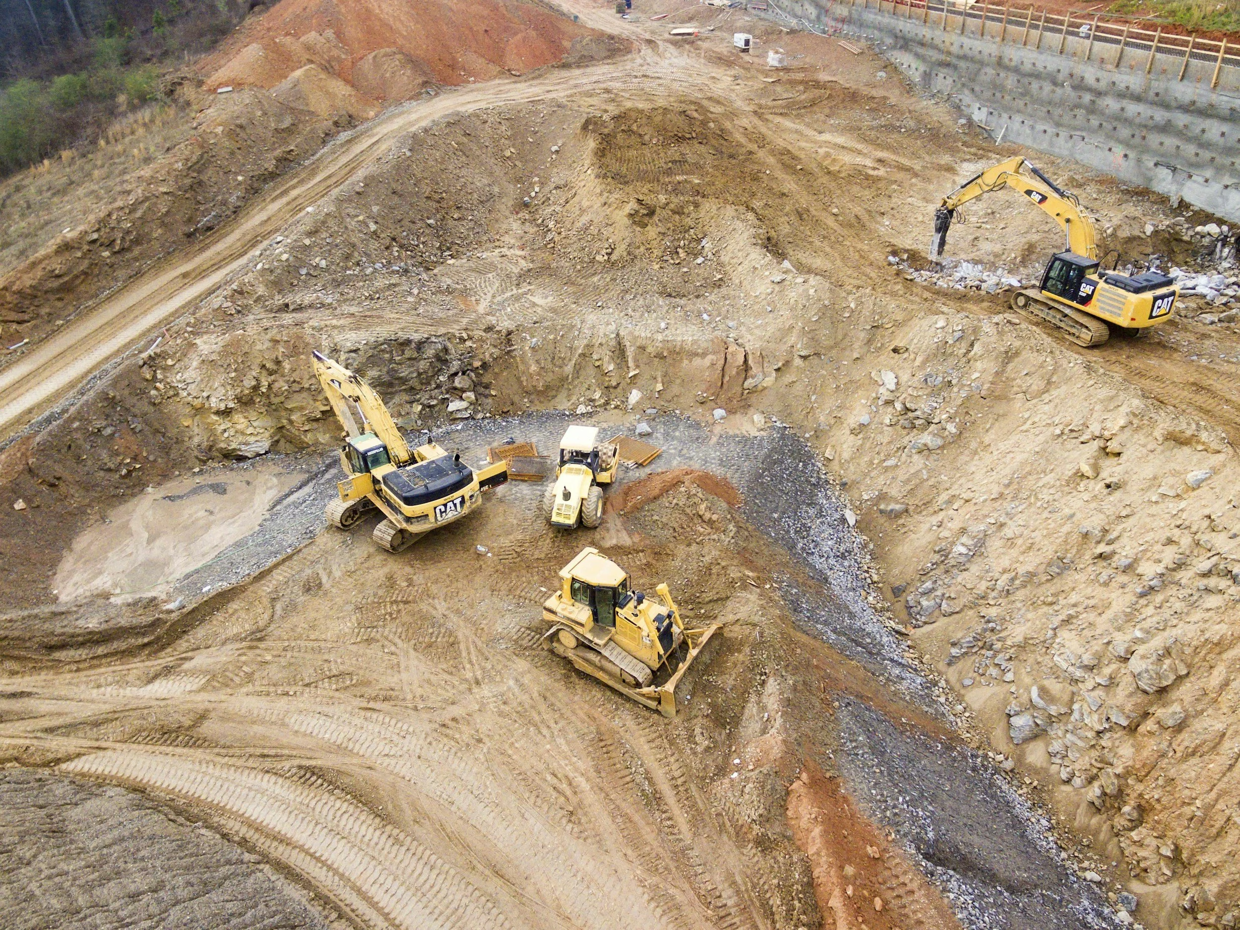Construction site with multiple excavators working on earth-moving and site preparation, with dirt piles and a retaining wall in the background.