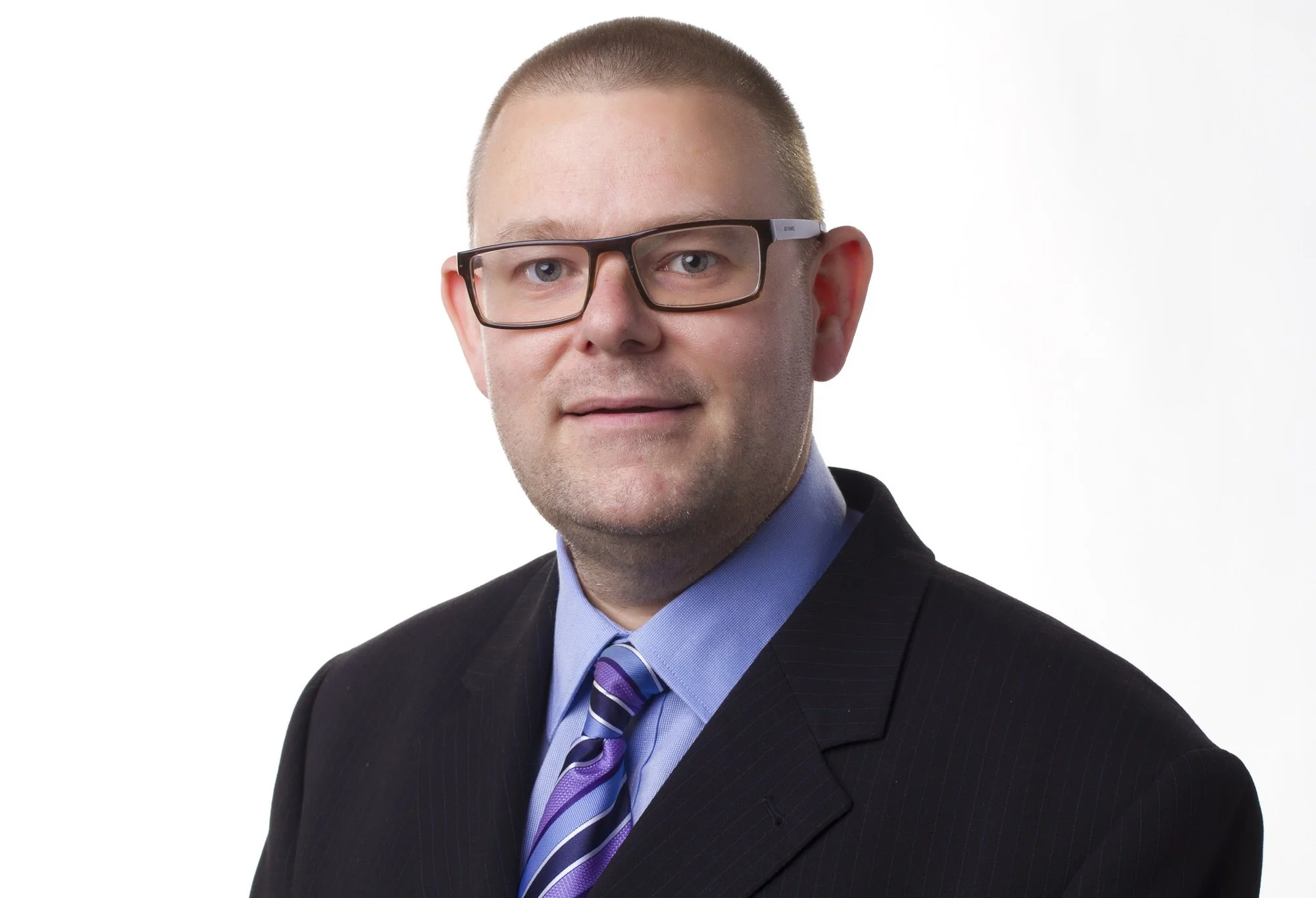 Professional headshot of a man with short hair, glasses, wearing a dark suit, light blue shirt, and a striped purple tie, against a white background.