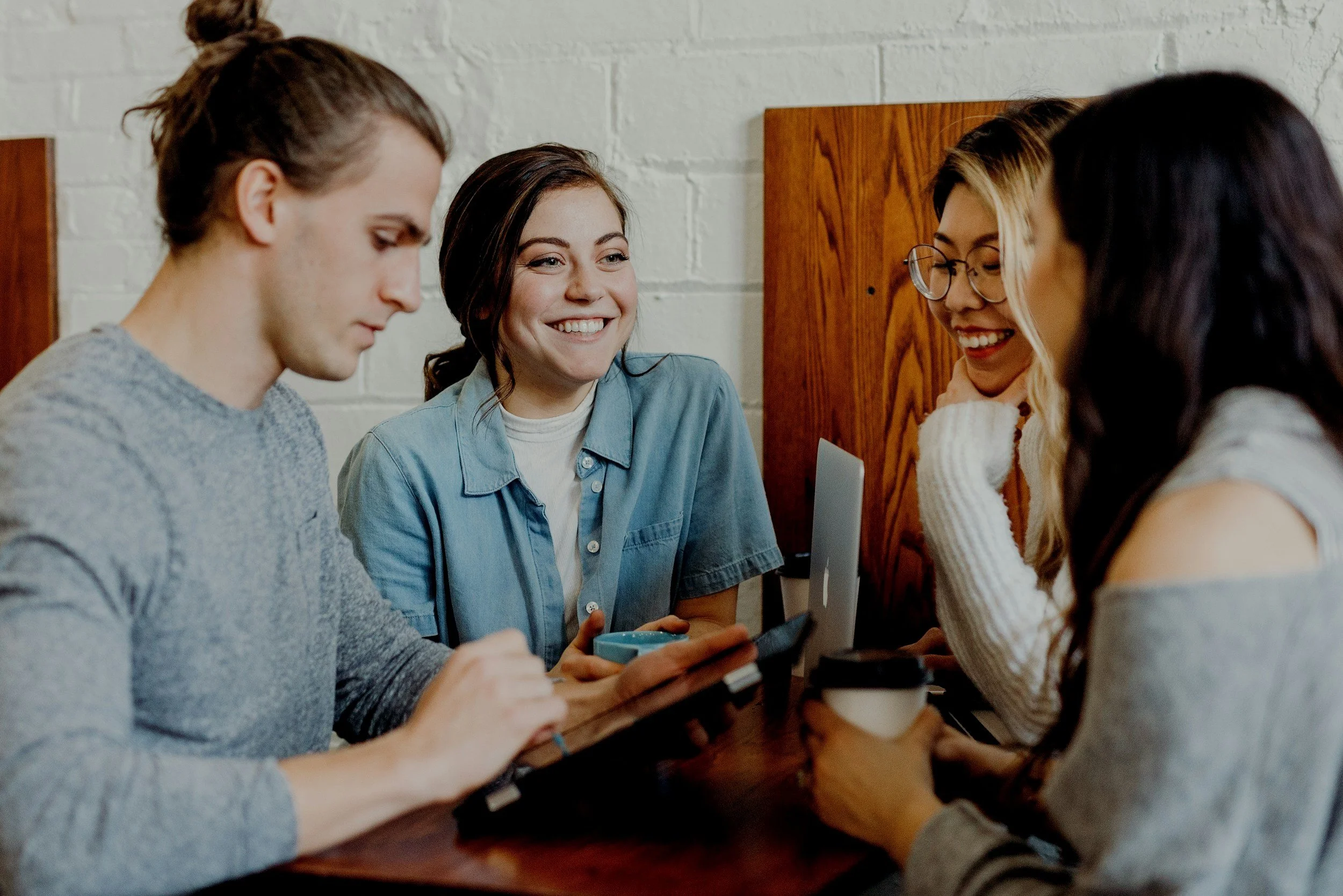 Four young adults sitting at a table in a casual setting, engaging with each other and using electronic devices, with a white brick wall in the background.