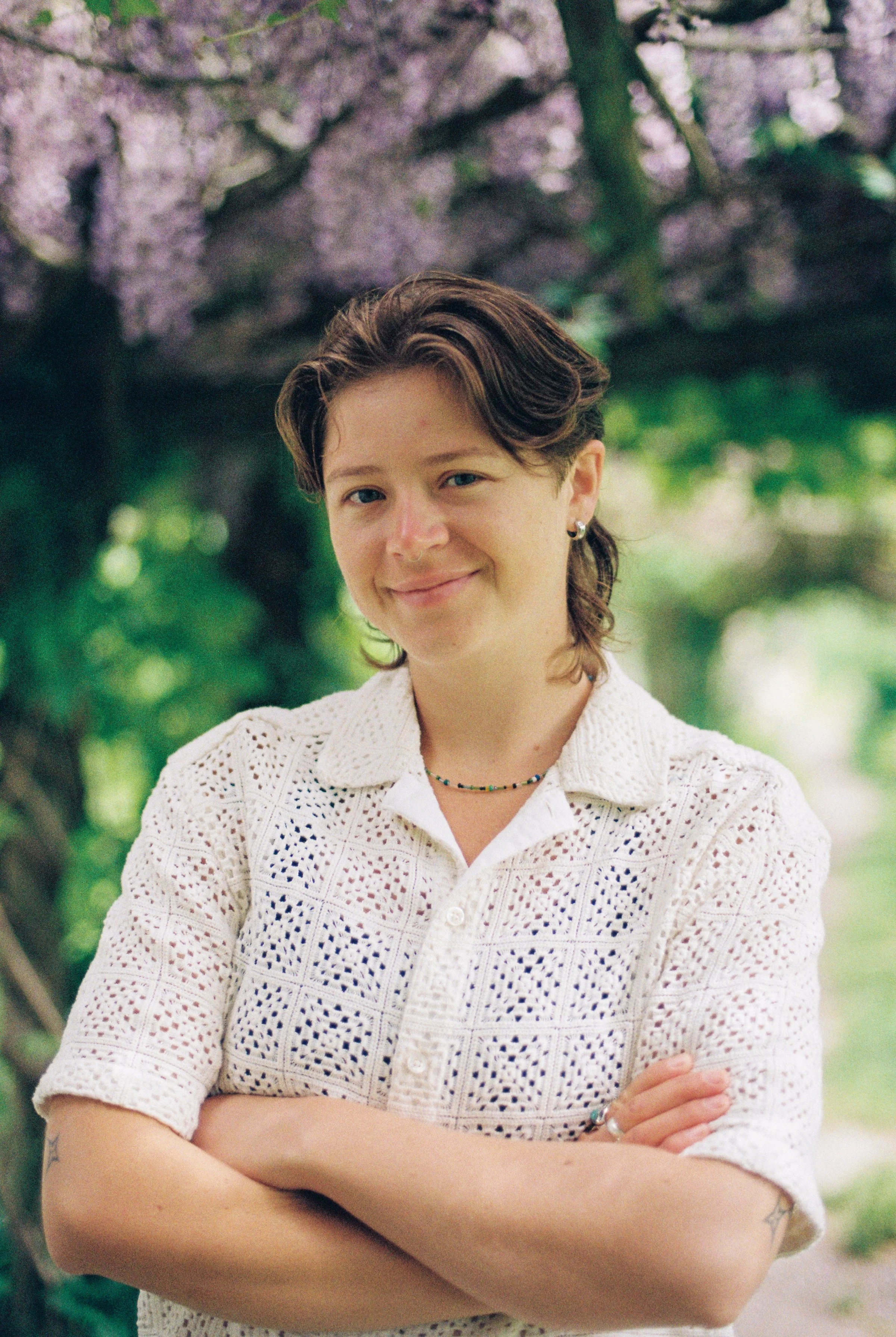 A person with short, brown hair and blue eyes, smiling with arms crossed, standing outdoors in front of a flowering tree with pink blossoms and green leaves.