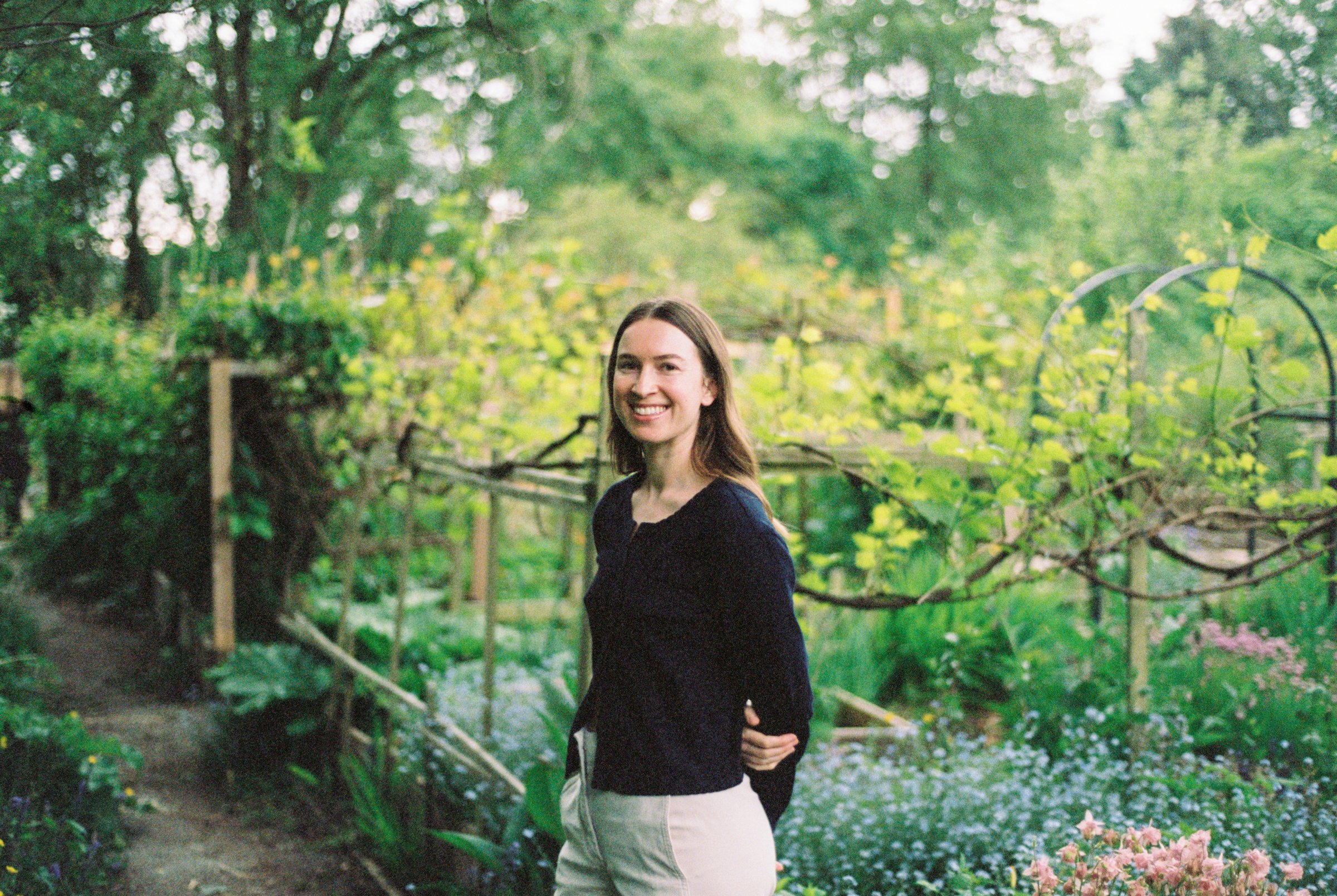 A woman with long brown hair wearing a dark sweater and light-colored pants, smiling and standing in a lush garden with green plants, trees, and colorful flowers.