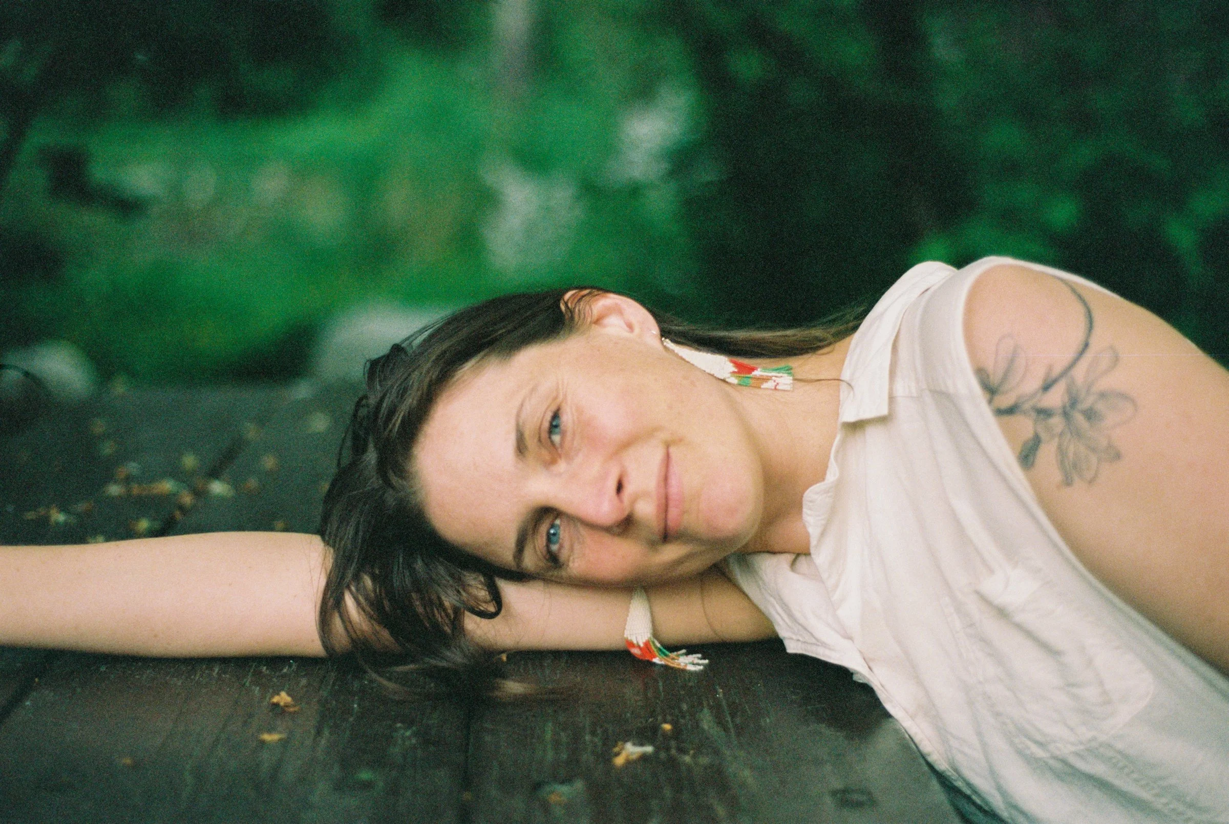 A woman with dark hair and blue eyes lying on a wooden surface, with a green blurred background, wearing a white sleeveless shirt and a colorful earring.