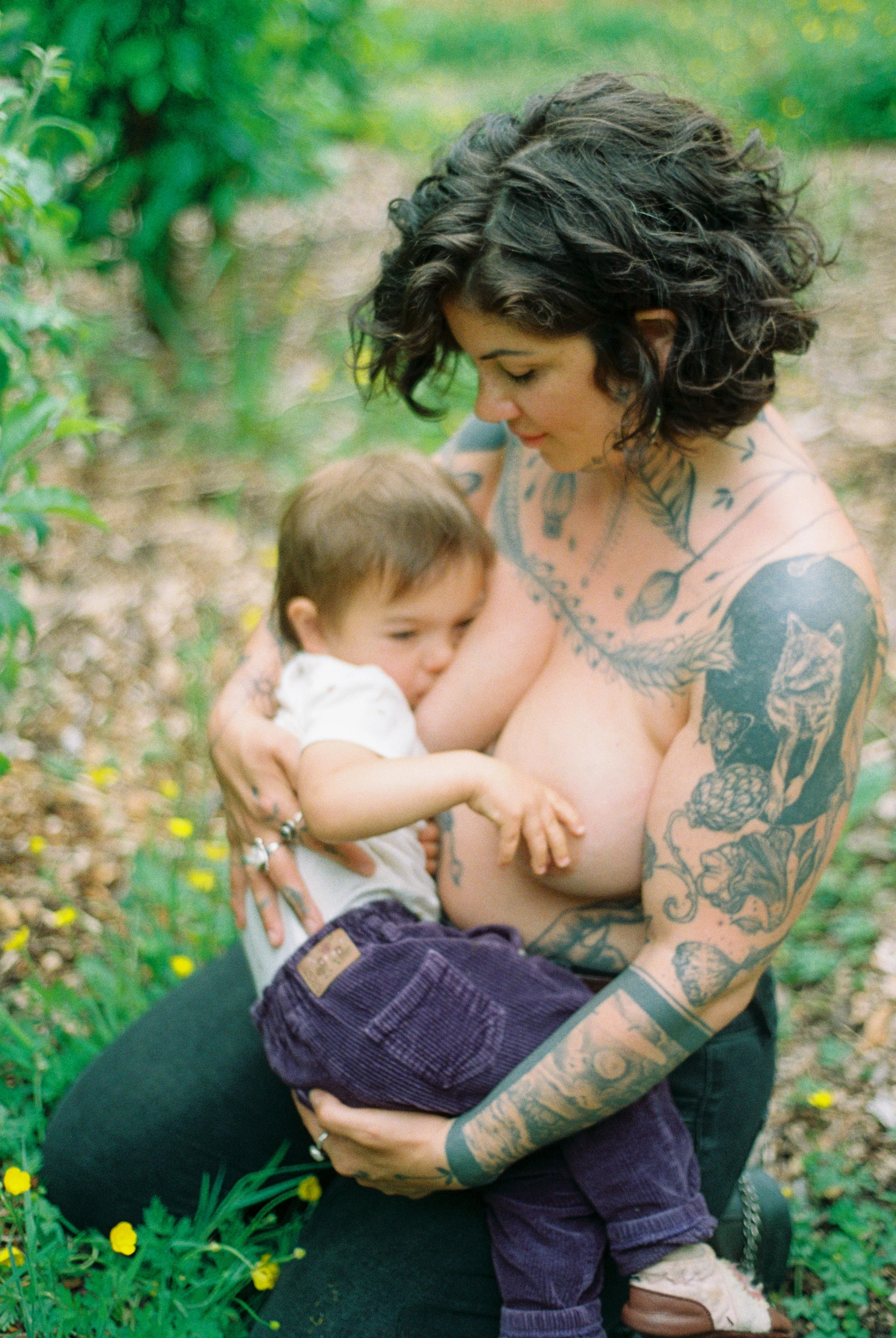 A woman with tattoos on her arms and chest holding a young child outdoors in a green, garden setting.