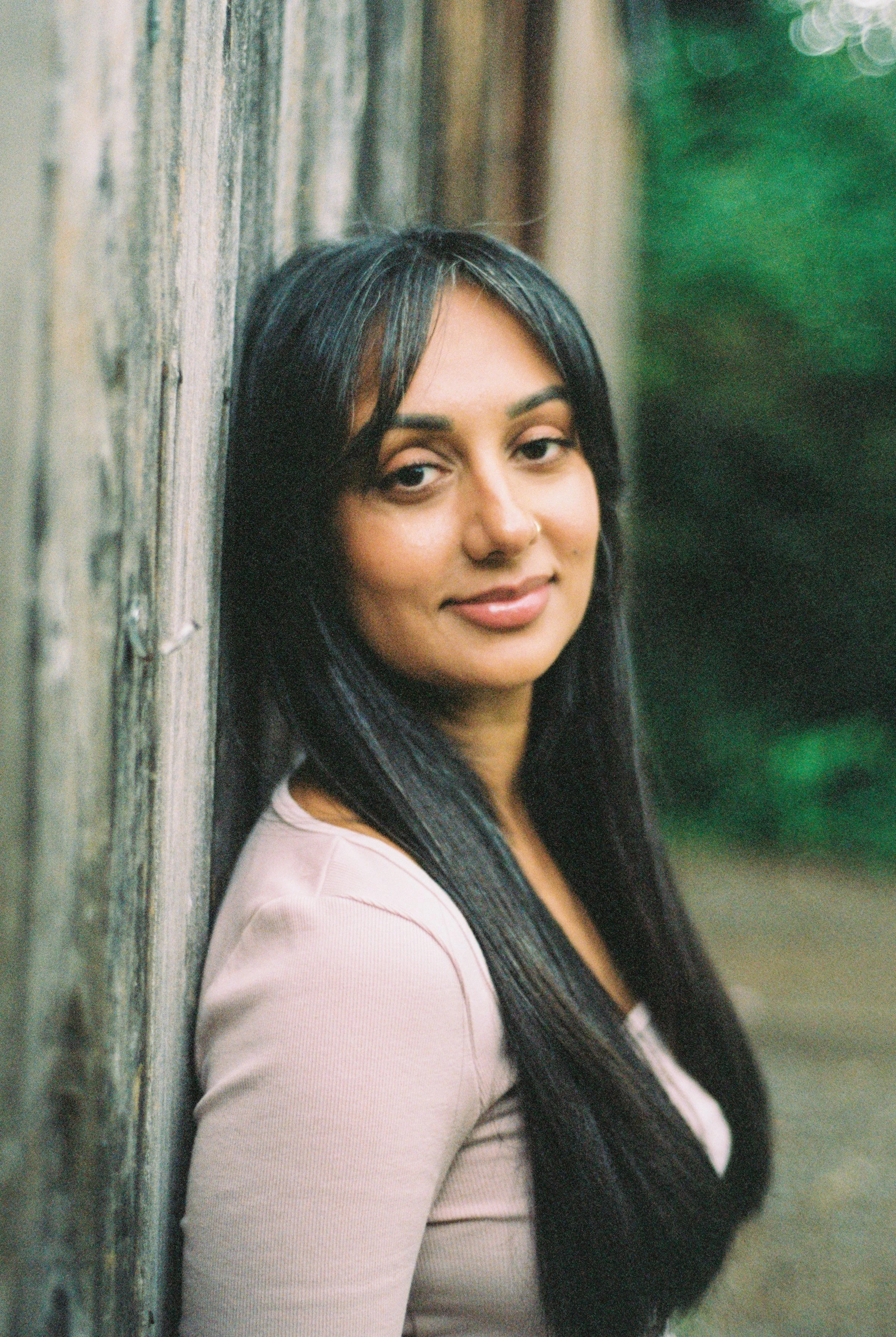 A woman with long, straight black hair and a nose ring, leaning against a wooden fence, looking at the camera, outdoors with greenery in the background.