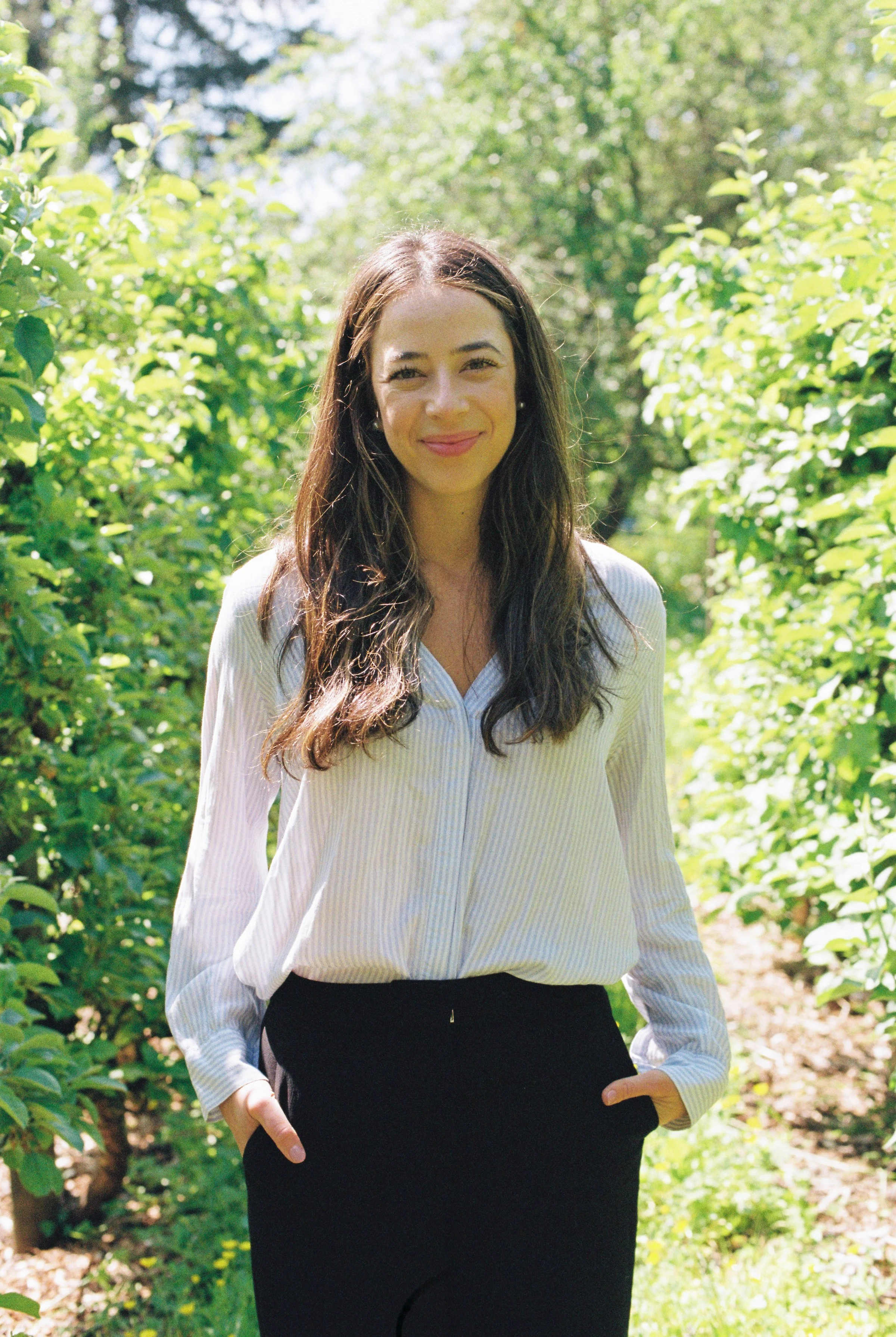 A young woman with long dark hair and a smile, standing outdoors among green bushes and trees with bright sunlight, wearing a white striped blouse and black pants.