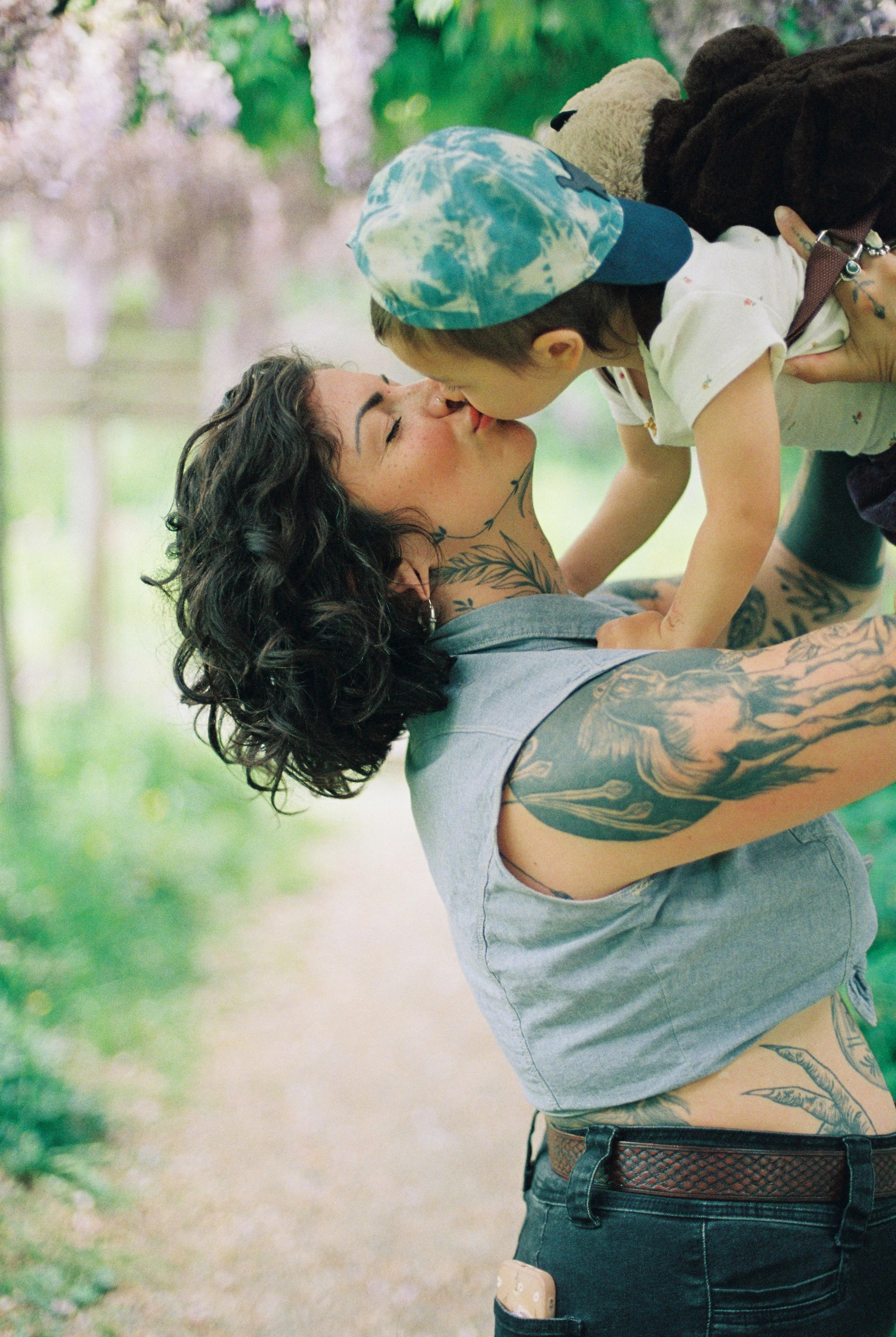 A woman with tattoos and curly hair kissing a young boy wearing a blue tie-dye cap outdoors.