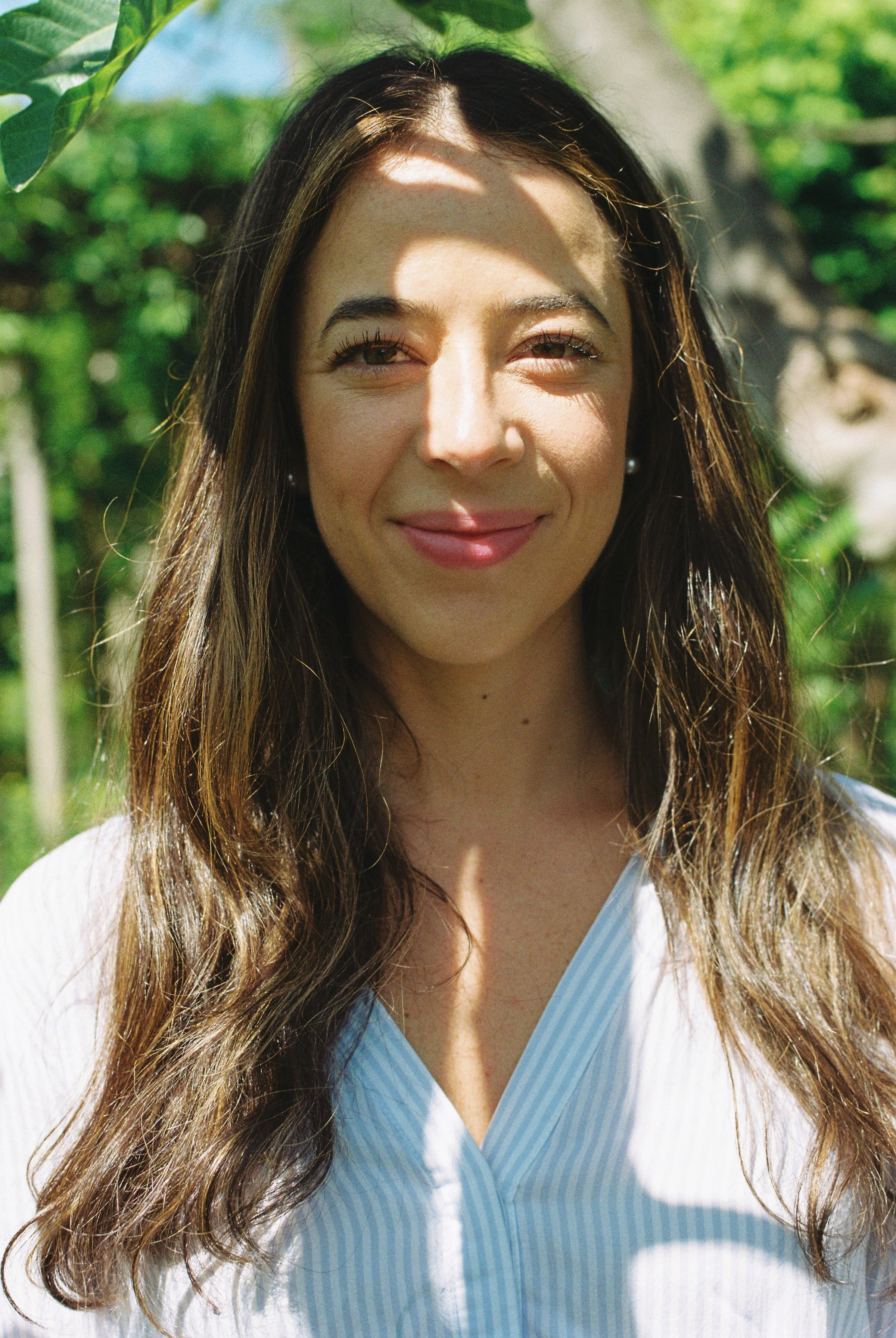 A woman with long brown hair smiling outdoors in a sunny environment with green foliage and trees in the background.