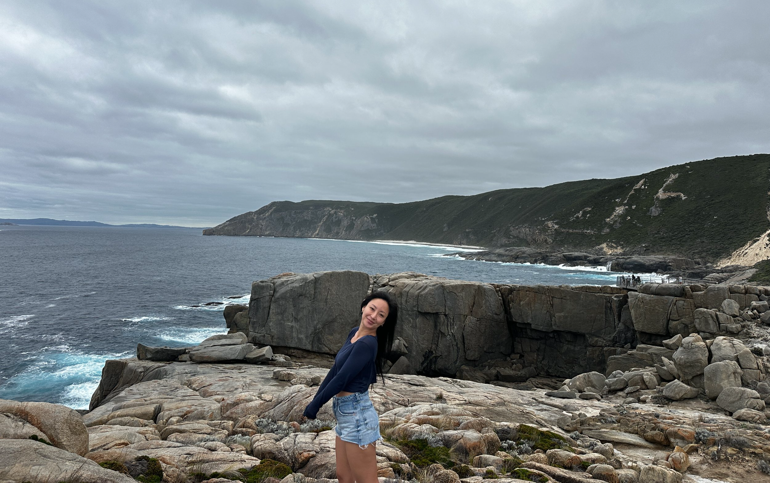 A woman stands on a rocky coastline with ocean waves and green hills in the background on a cloudy day.