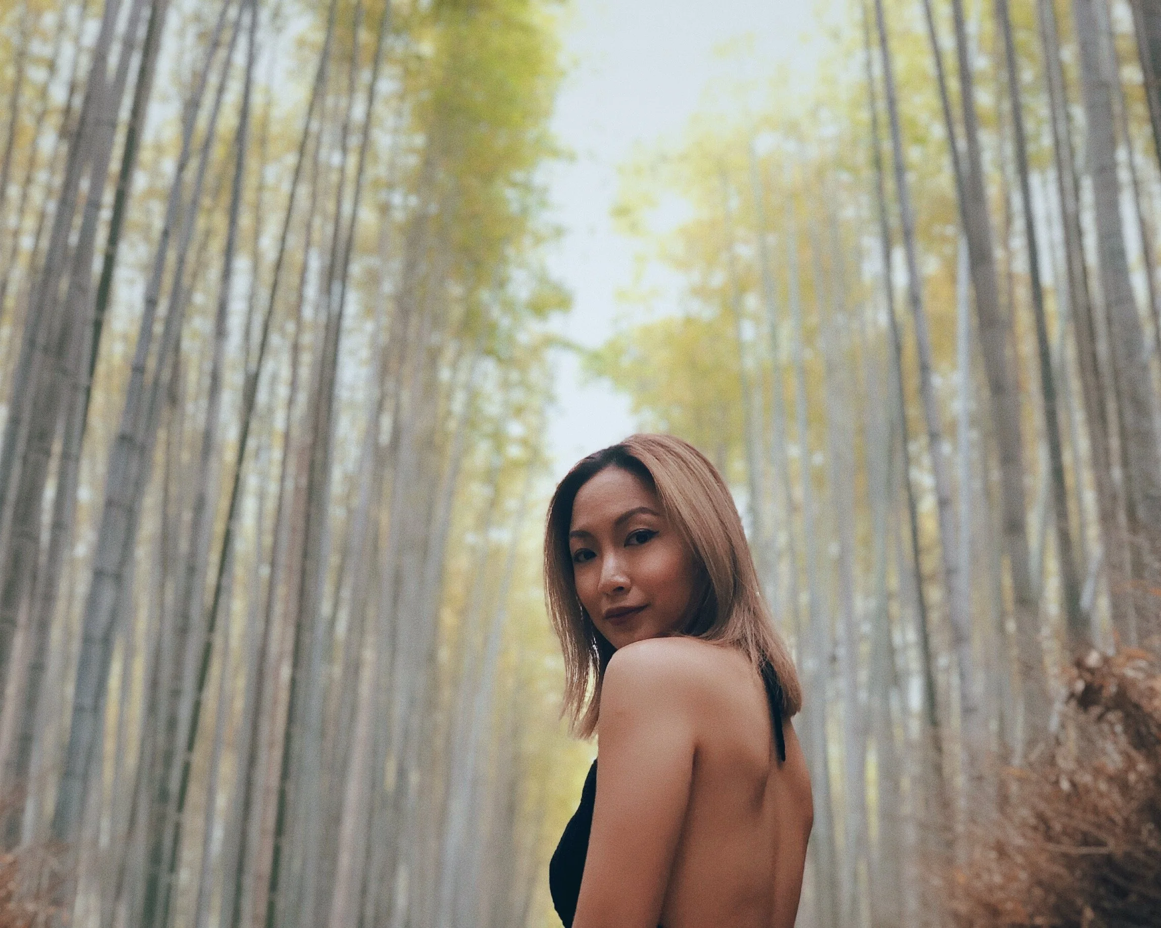A woman with shoulder-length brown hair and wearing a black dress stands in a bamboo forest during daytime, with tall bamboo stalks on either side and sunlight filtering through the leaves.