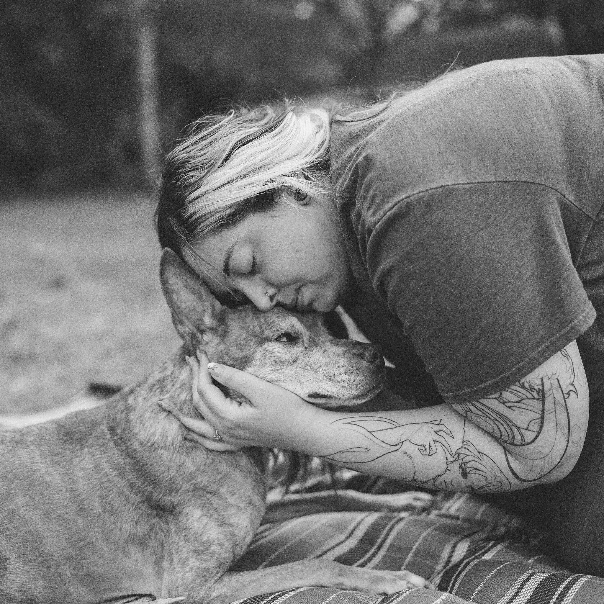 A woman with short hair and tattoos is laying on her side, pressing her forehead and nose gently to a dog's head. The dog is resting with closed eyes. It appears to be a peaceful moment of affection and bonding outdoors.