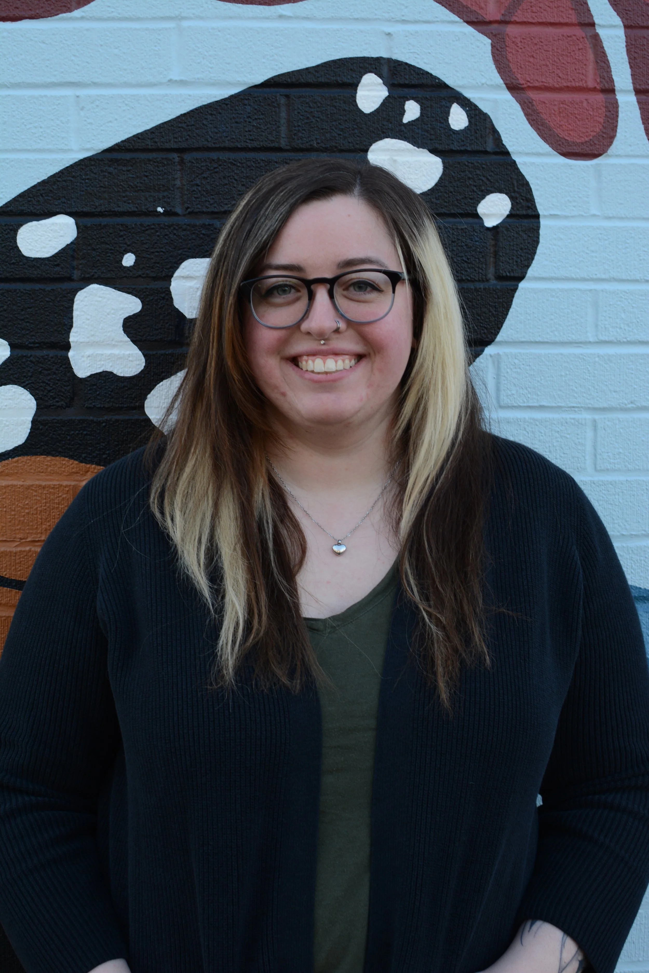 A young woman with glasses and light-colored hair standing in front of a colorful mural painted on a brick wall.
