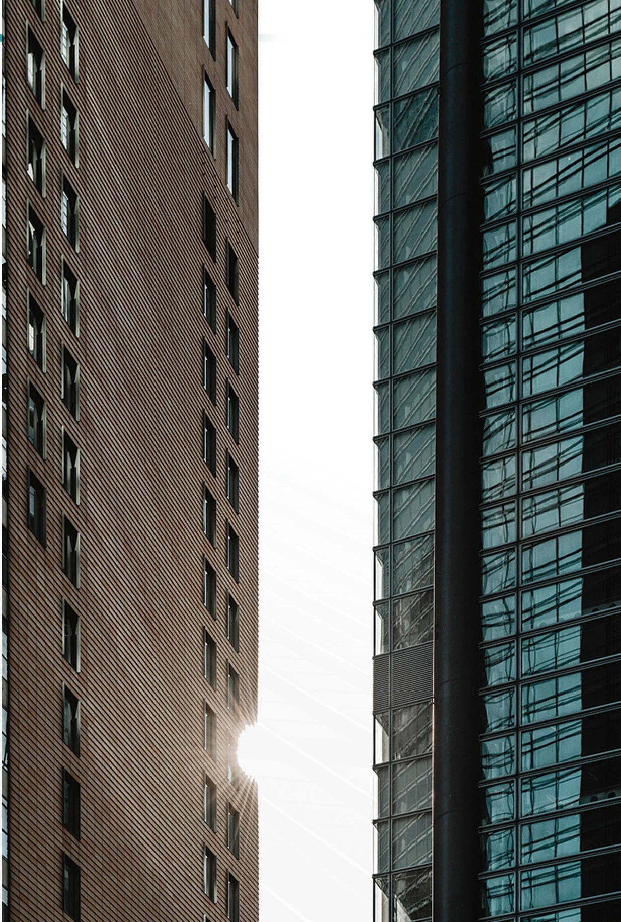 The image shows two tall modern buildings with the sky in between. The building on the left has a brown exterior with a repetitive pattern of rectangular windows, while the building on the right has a glass exterior reflecting the surroundings.
