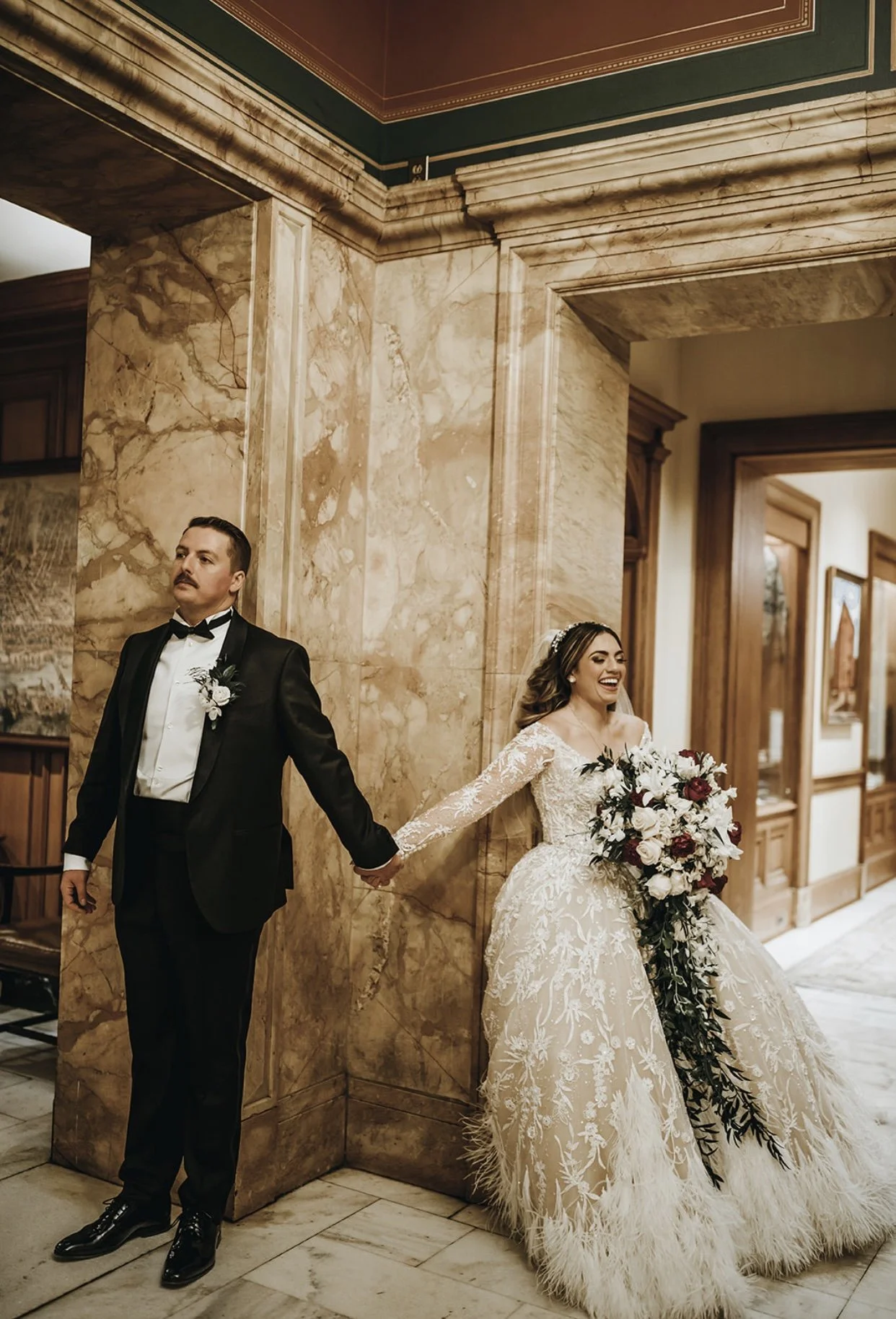 A bride and groom holding hands behind a marble column in an elegant interior, with the bride smiling and the groom looking serious.