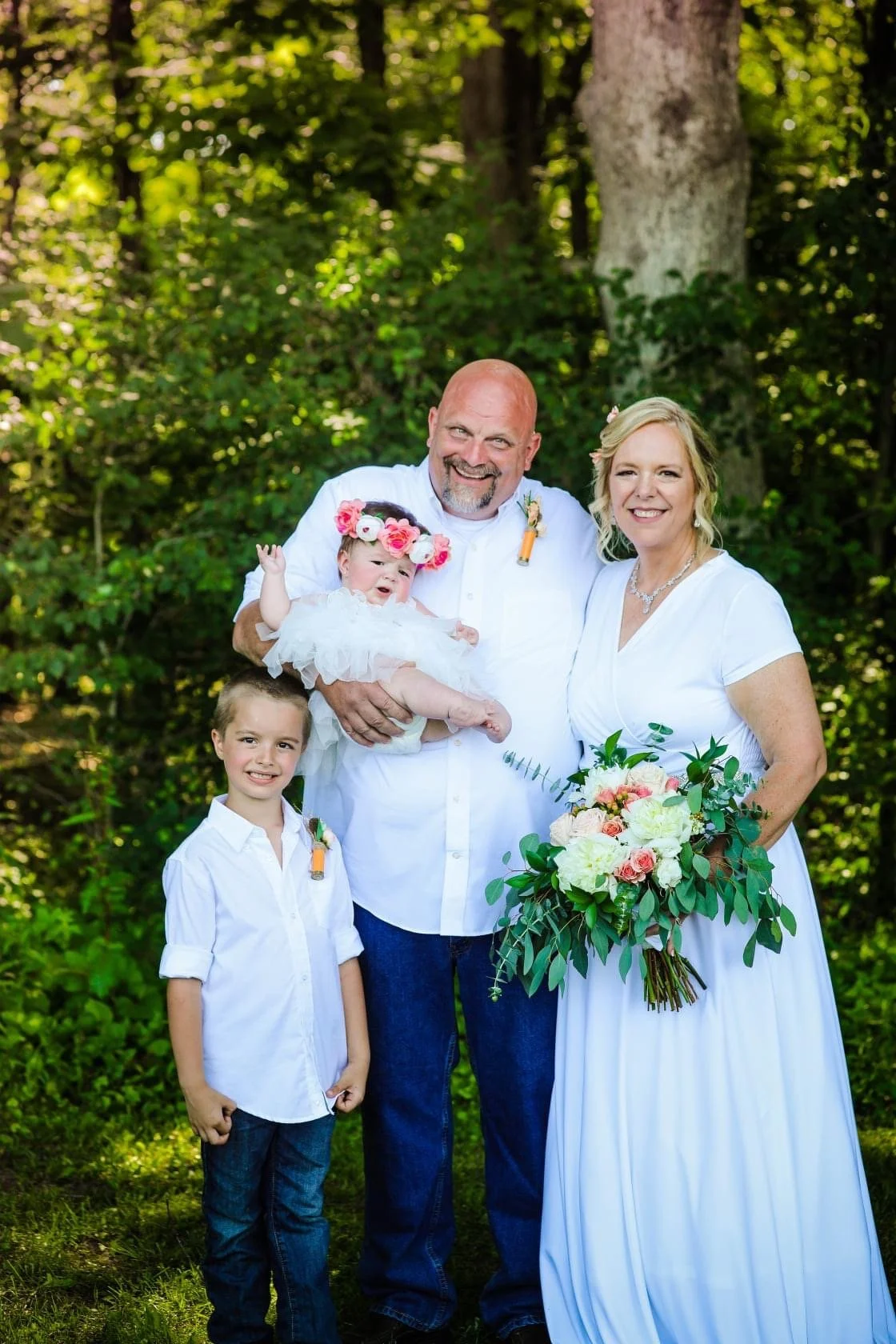A family of four dressed in white, standing outdoors in a wooded area, celebrating a special occasion. The woman is holding a bouquet of flowers, and the man is holding a baby girl wearing a flower crown. A young boy stands beside them, all smiling.