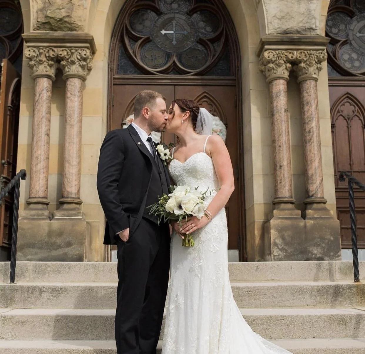 A bride and groom sharing a kiss on the steps outside a church. The bride is in a white wedding dress holding a bouquet of white roses, and the groom is in a black suit with a boutonniere.
