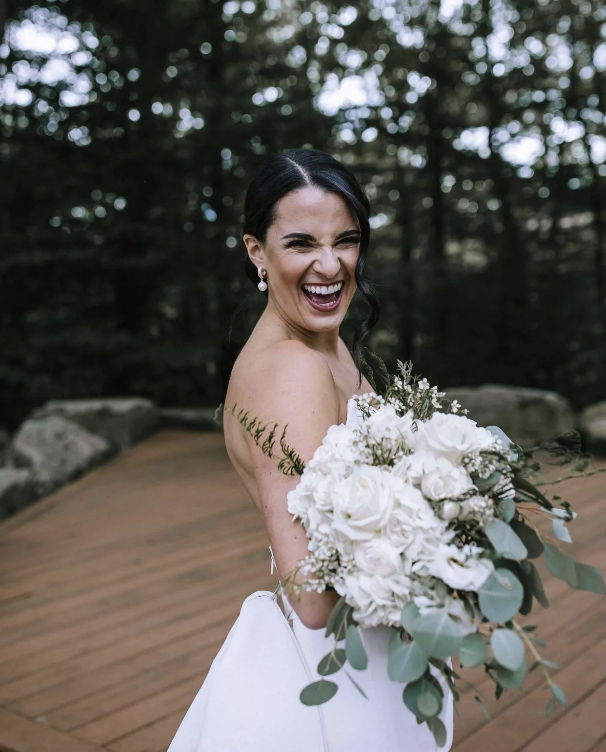 A woman in a wedding dress holding a bouquet of white flowers and green foliage, smiling and laughing outdoors with trees in the background.