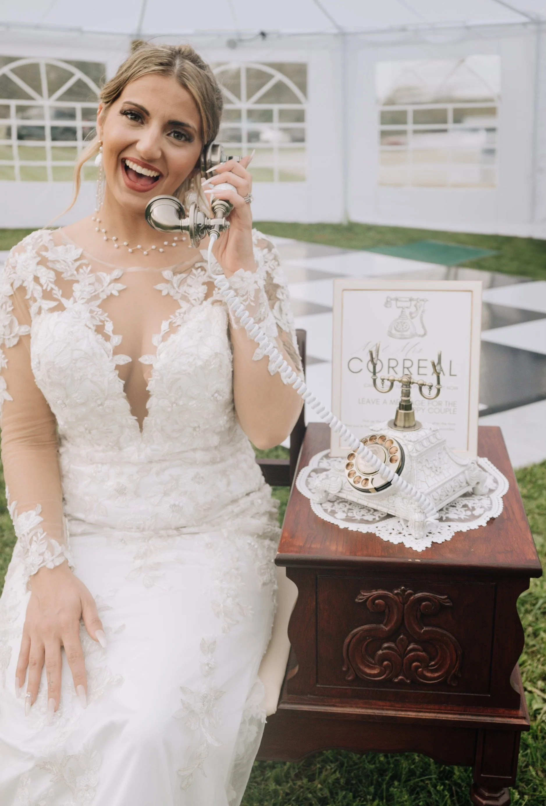 Bride in a lace wedding dress talking on a vintage telephone at a wedding reception tent.