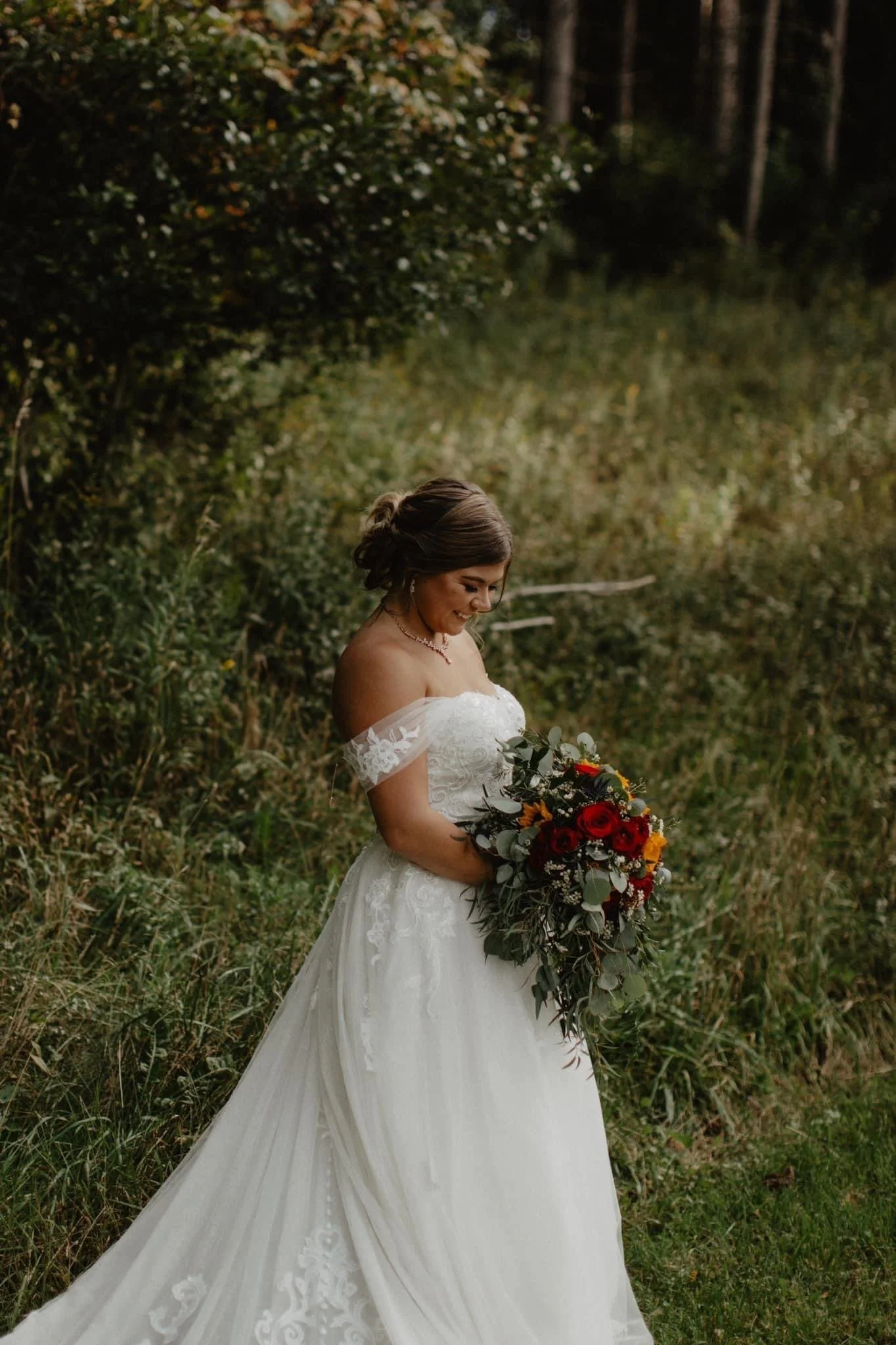 Brunette bride in white wedding dress holding a colorful bouquet, standing outdoors in a green, wooded area.