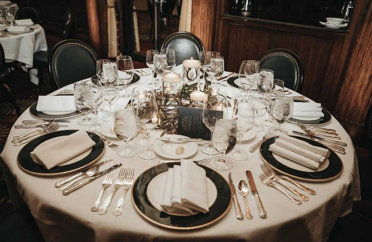 Elegant round table set for a formal dinner with black and gold plates, white napkins, multiple wine glasses, candles, and decorative leaves, in a dark wood-paneled room.