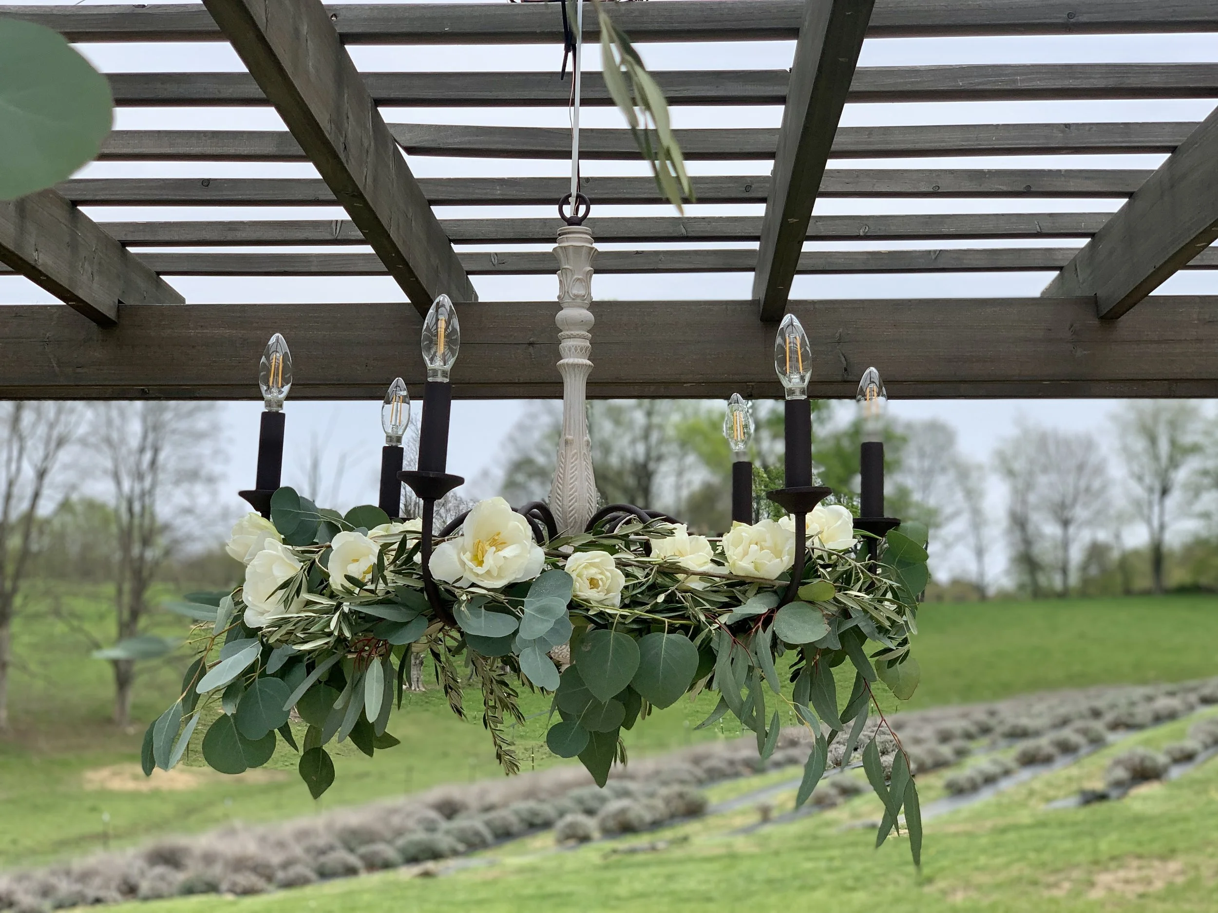 Chandelier with white flowers and green leaves hanging under a wooden pergola in an outdoor garden.